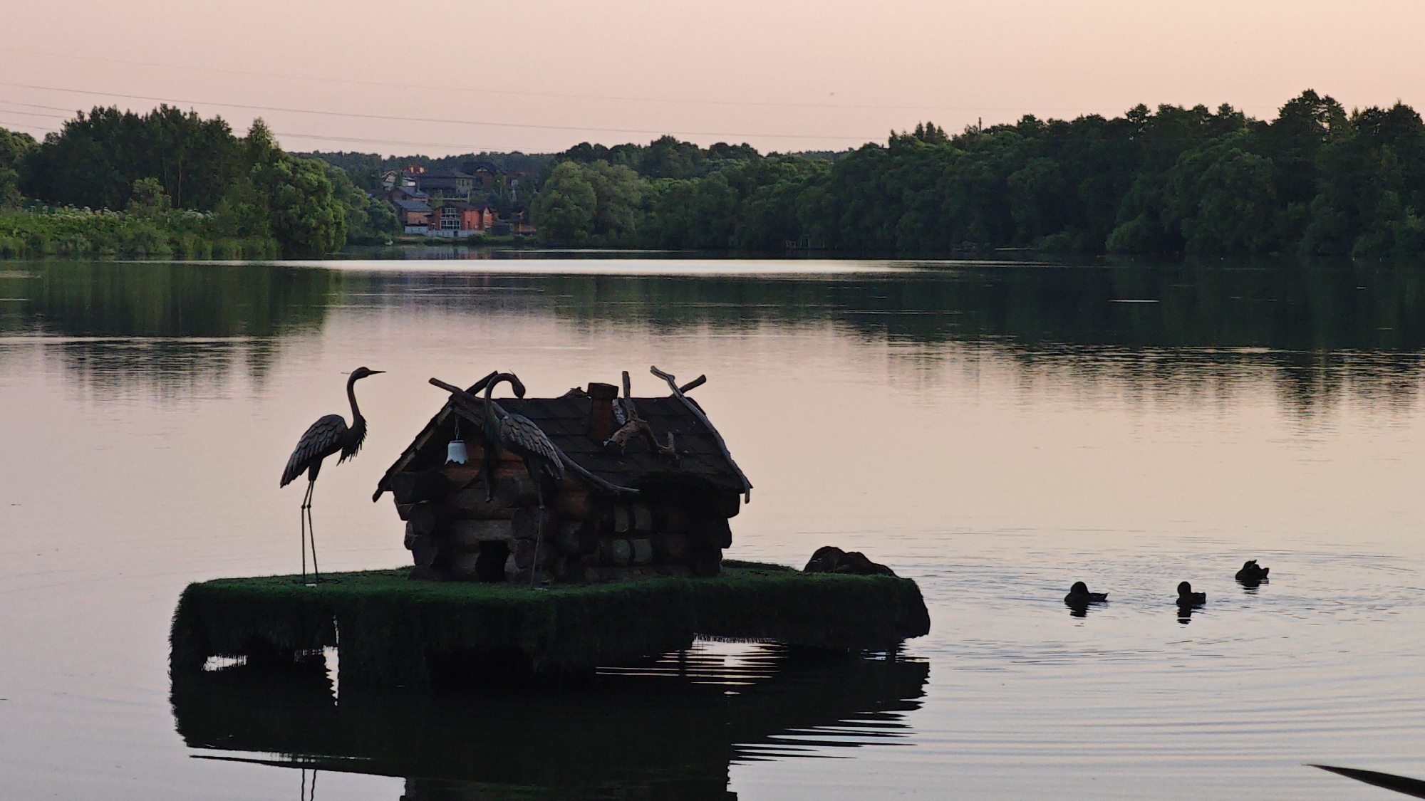 The image depicts a tranquil lake scene at dusk, possibly in or near Aleksandrovo, Russia. In the foreground, a small, intricately crafted wooden house sits on a floating island covered in grass. A silhouette of a heron stands beside the house, its long legs planted firmly on the island. Two ducks are visible swimming in the calm water to the right of the island, with a third duck further to the right. The background features a dense line of trees and a few houses nestled amongst them, with a pale pink sky overhead. The water reflects the trees and the sky, creating a serene atmosphere.