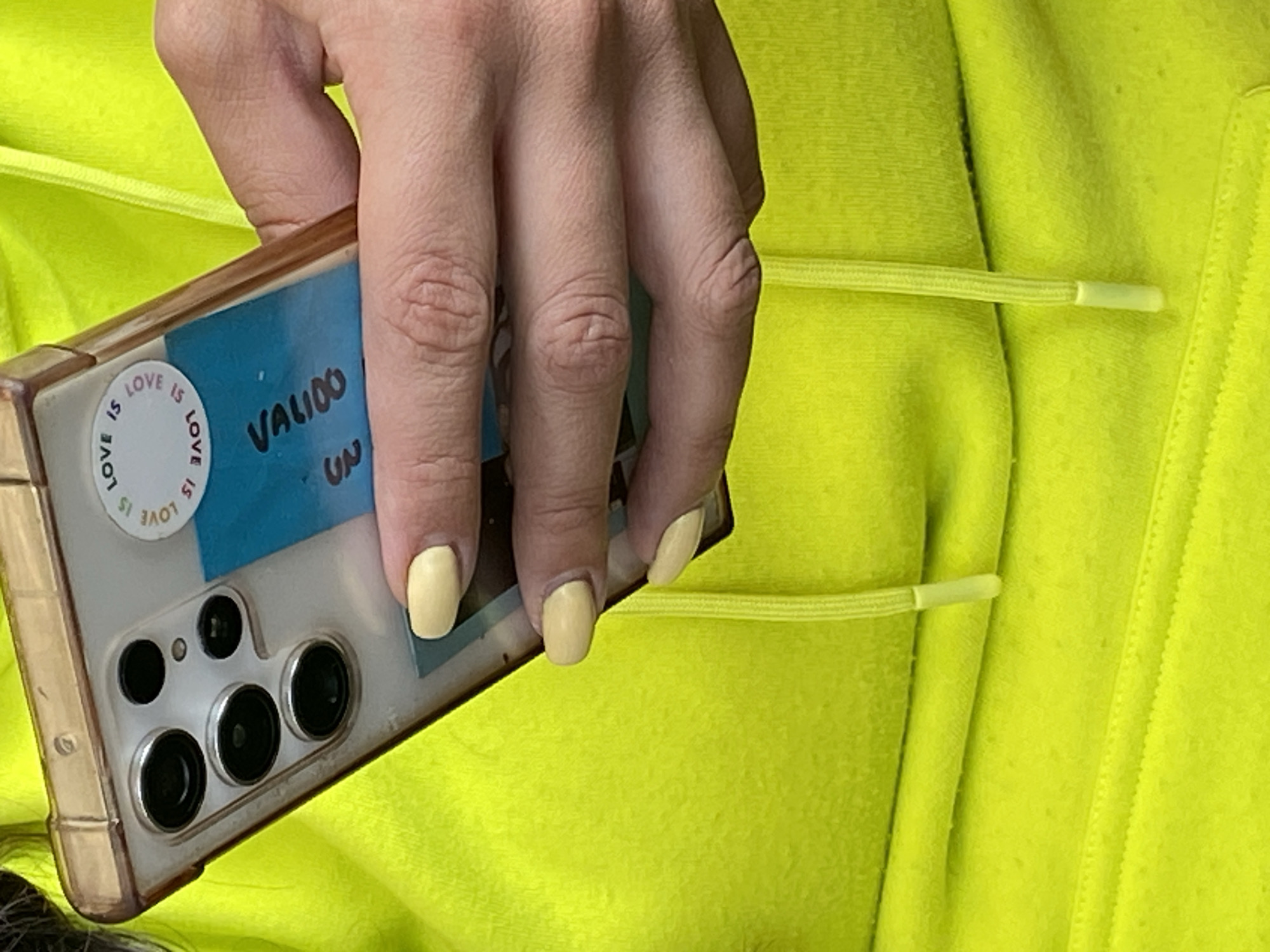 A person's hand holds a smartphone in a transparent case against a bright yellow garment, likely a hoodie. The smartphone case is decorated with two stickers: a circular one stating "LOVE IS LOVE" and a blue rectangular one with "VALIDO" written on it. The phone's multi-lens camera system is visible. The person's fingernails are painted a pale yellow color. There's no information given in the image that allows me to place it in the city of Puebla, Mexico.
