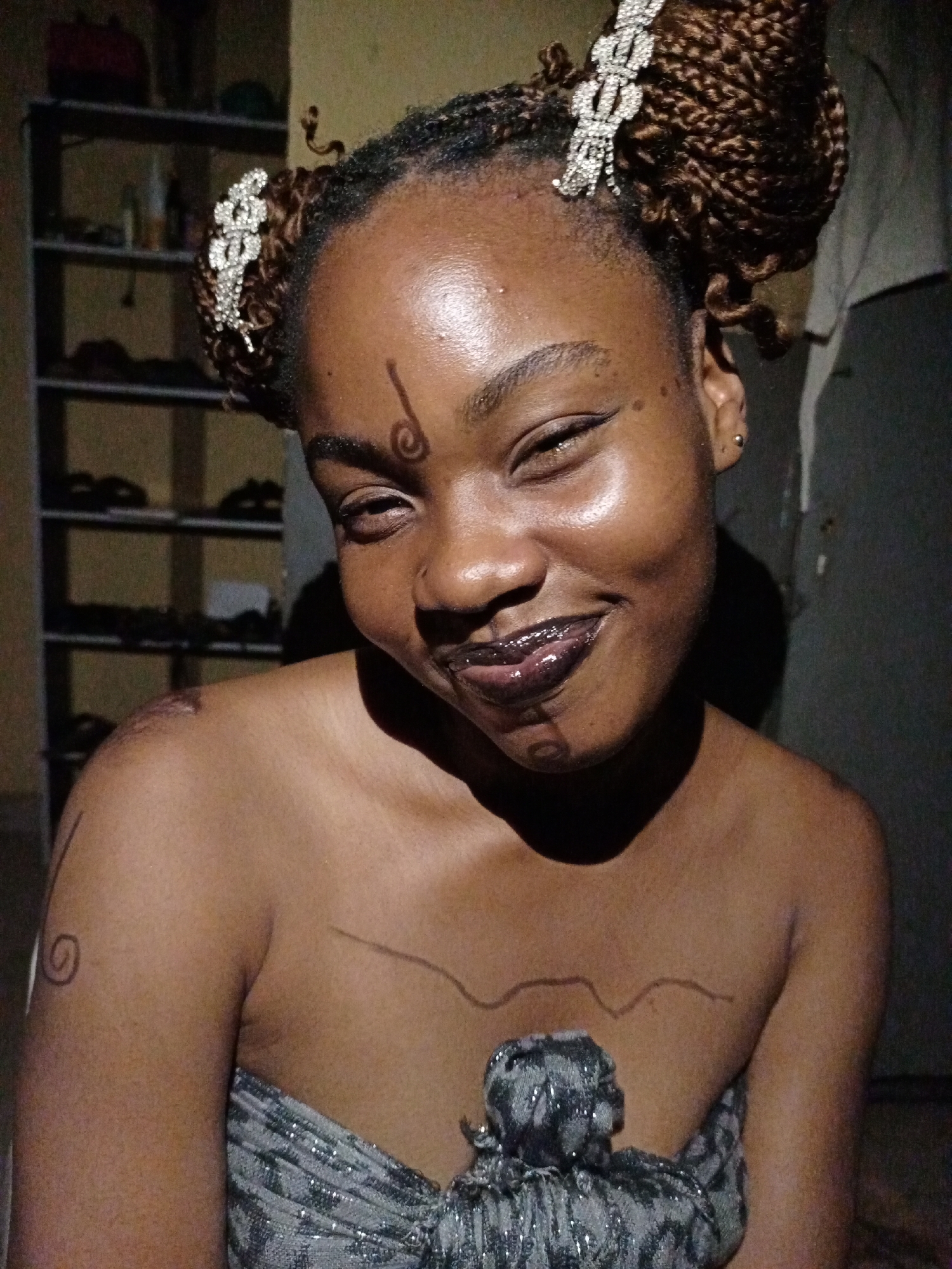 A young Black woman is depicted in a close-up, dimly lit portrait, smiling broadly with her eyes squinted. She has dark skin and her hair, styled in brown box braids, is pulled up into two buns adorned with elaborate, sparkling hair clips that resemble ornate bows. She wears dark eyeliner and dark, glossy lipstick or lip gloss.
Distinctive body art is drawn on her skin: a vertical line terminating in a spiral is centered on her forehead, resembling a stylized musical note. A subtle vertical line is on her left cheek. On her right shoulder (viewer's left), a spiral is drawn, and a wavy line extends across her upper chest. Another vertical line is visible on her left upper arm (viewer's right).
She is clad in a strapless garment, possibly a wrap top, which appears to be a greyish, metallic, and sparkling fabric. The background is dark and features a multi-tiered shelf unit with indistinct objects, suggesting an indoor domestic setting with low light, possibly at night. The woman's expression conveys joy and happiness. The image depicts a moment of personal adornment or cultural expression, set in Keffi, Nigeria. No visible text is present.