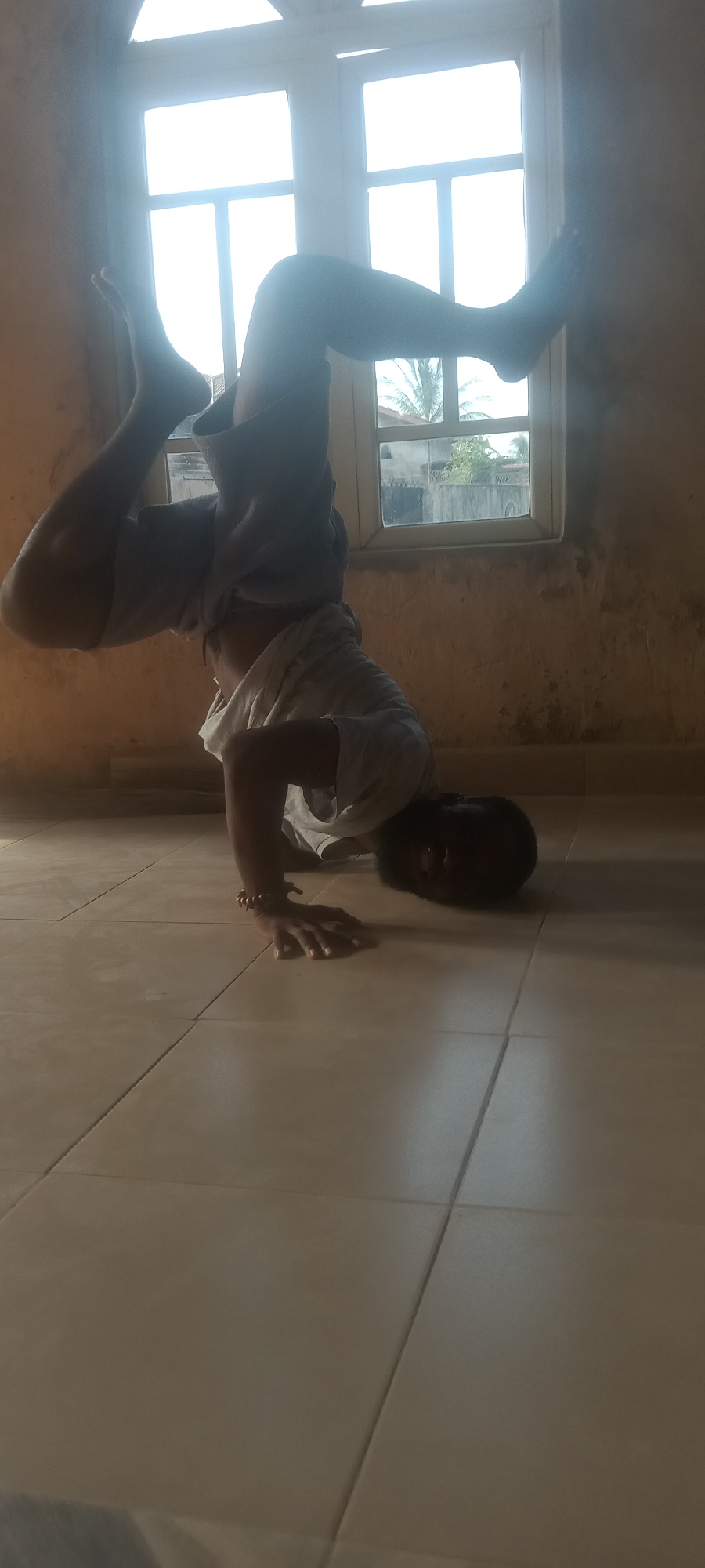 A person is performing a headstand indoors. The individual is positioned on a tiled floor with their head and hands supporting their weight. The person is wearing shorts and a light-colored t-shirt. A window with a grid pattern is visible in the background, revealing a glimpse of the outside, with hints of trees. Based on the request, this is in Bende, Nigeria. The room appears to be sparsely furnished, with the tiled floor and a painted wall.