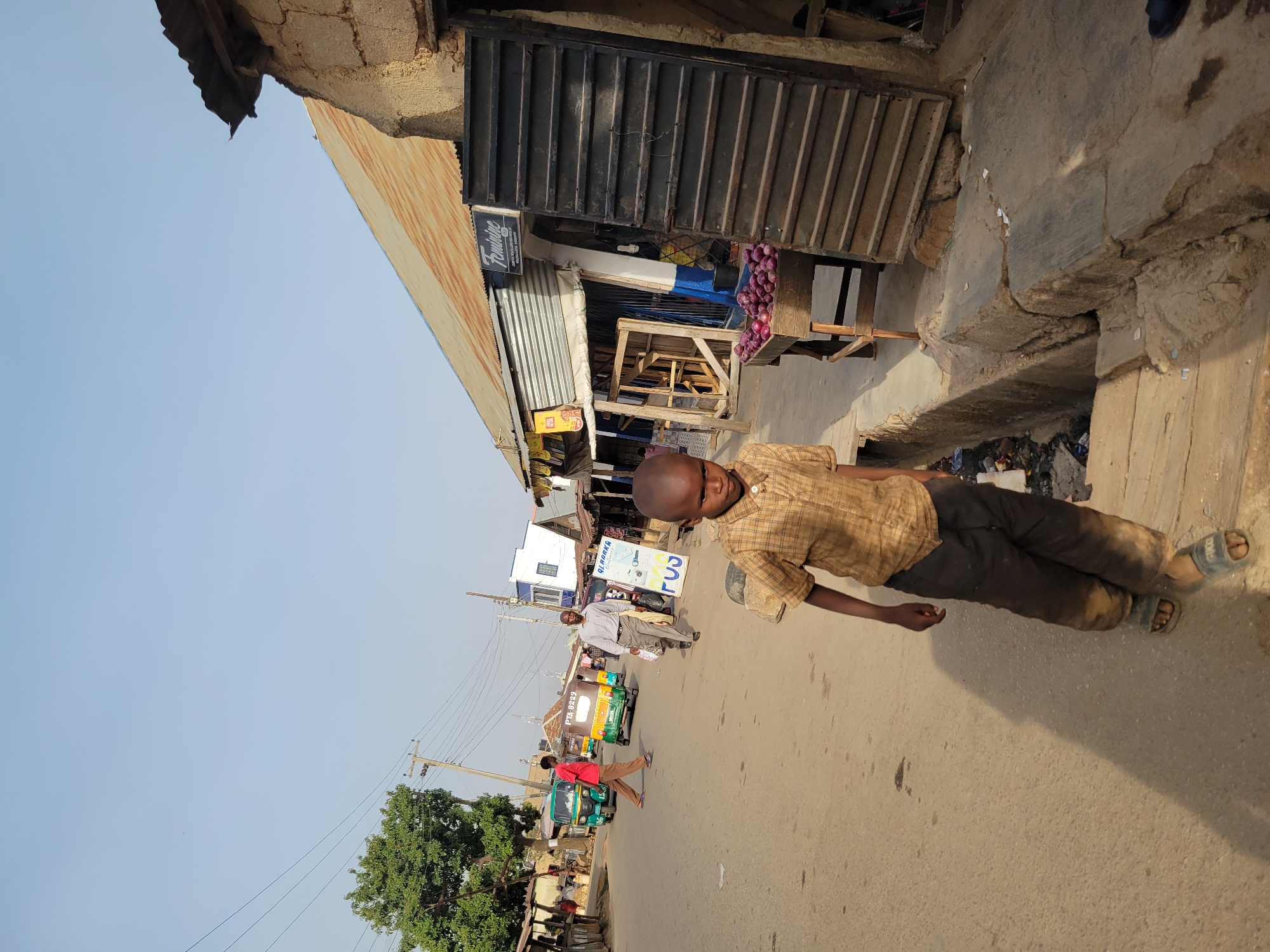 A young boy wearing a plaid shirt and dark pants walks along a street in Jos, Nigeria. He is on the right side of the image, near the edge of a building. In the background, there are people, trees, tuk-tuks, and other buildings, suggesting a street scene in a city.
