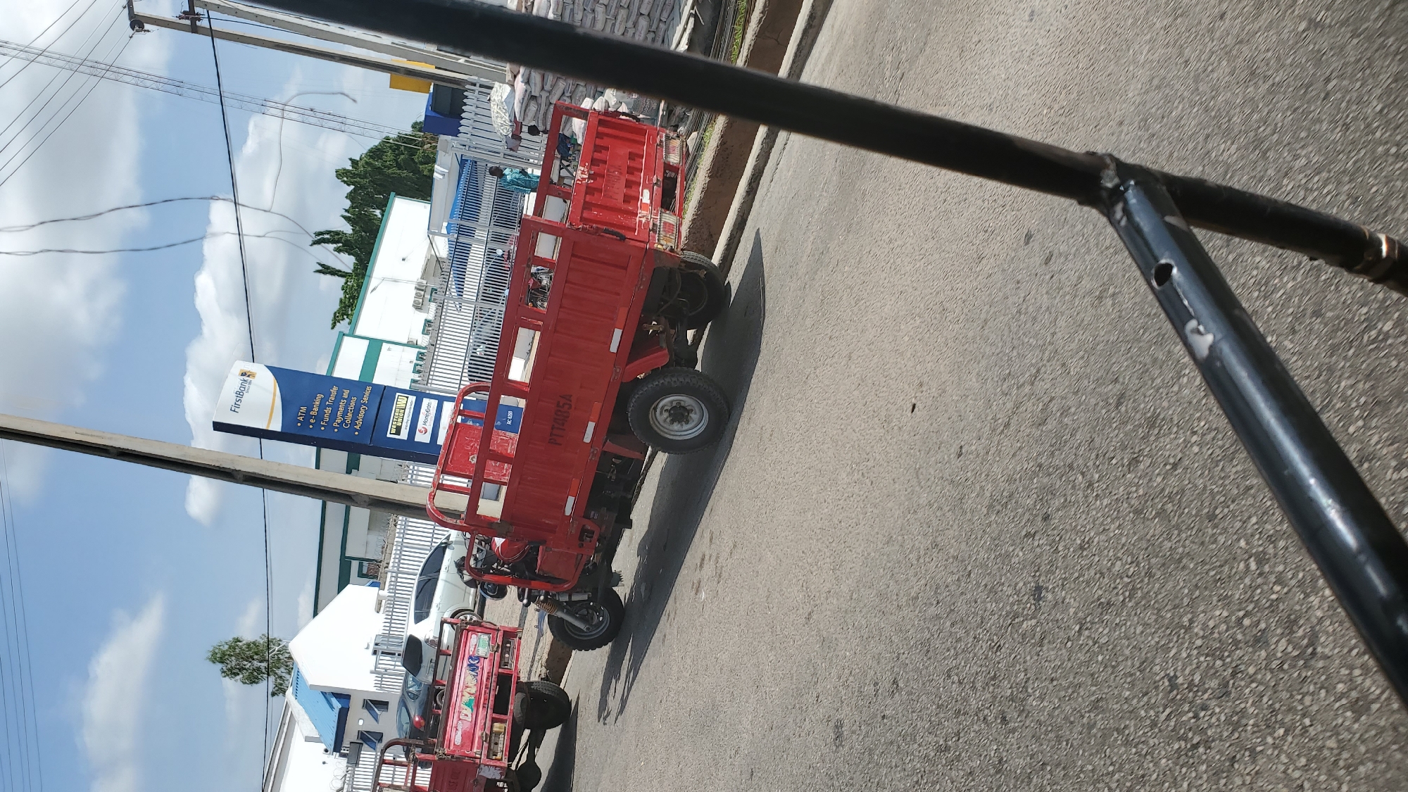 In the city of Jalingo, Nigeria, a red motorized tricycle, identified by the license plate "PTT485A", is parked on an asphalt road. The tricycle has a flatbed cargo area, typical for transporting goods. In the background, a building with a distinctive white and blue facade, bearing the "FirstBank" logo, suggests a financial institution. A metal pole, possibly a utility or street light pole, is prominently positioned in the foreground. Several white sacks, likely containing cement or similar materials, are stacked next to the building, indicating commercial activity or construction in the area. The sky is partly cloudy.