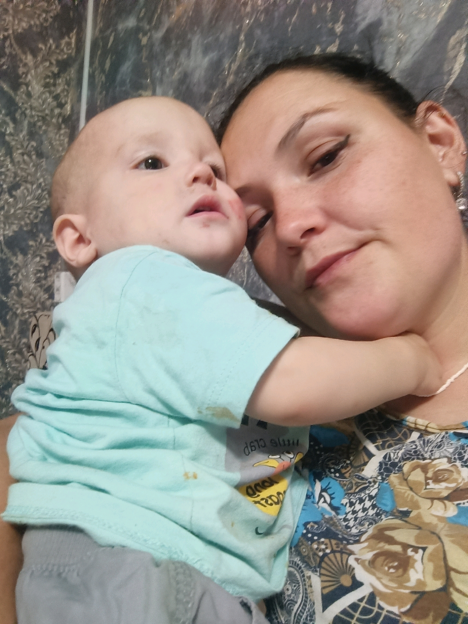 A woman and a baby are captured in a close-up selfie. The woman, with dark hair pulled back, is on the right side of the frame, her face tilted towards the camera. She is wearing a blue and floral patterned top. A baby, positioned on the left, is being held by the woman, with its head turned away from the camera. The baby is wearing a light blue t-shirt and grey bottoms. The background appears to be an interior wall with a patterned design. The image's lighting suggests it was taken indoors. There is no discernible information about the location being Karsha, Russia.