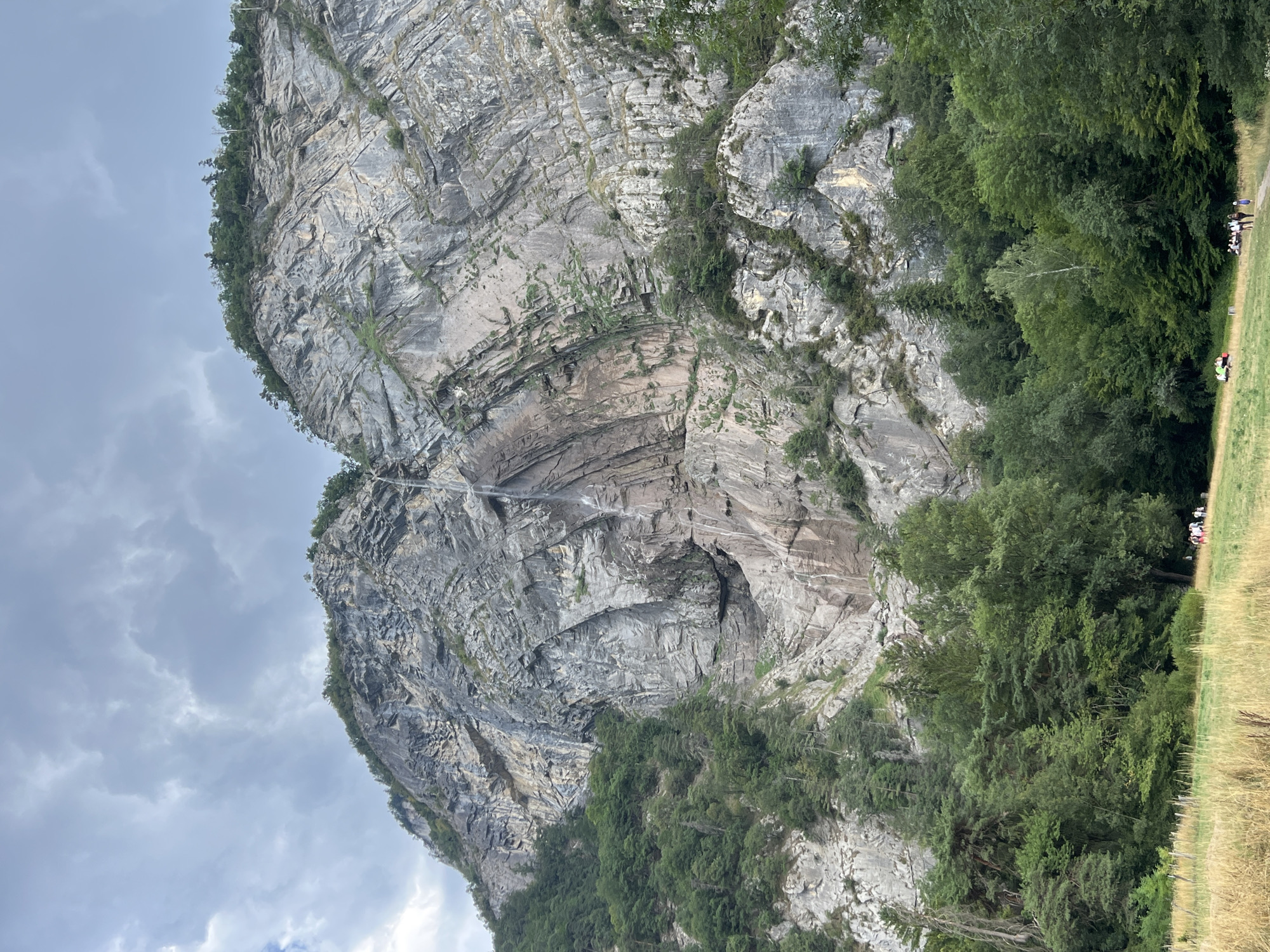 A dramatic waterfall cascades down a sheer, rugged mountain face, framed by lush green trees and a cloudy sky. The mountain is composed of layered rock, showing signs of erosion and natural formations. At the base of the mountain, a dense forest transitions to a grassy field where a small group of people are gathered. The field is dotted with dry grass and a few scattered trees. A dirt path winds through the field, leading towards the gathering of individuals who appear to be observing the waterfall or the scenery. The overall scene depicts a natural landscape with human presence. This appears to be the waterfall of Vergioz, located in the vicinity of Sallanches, France.