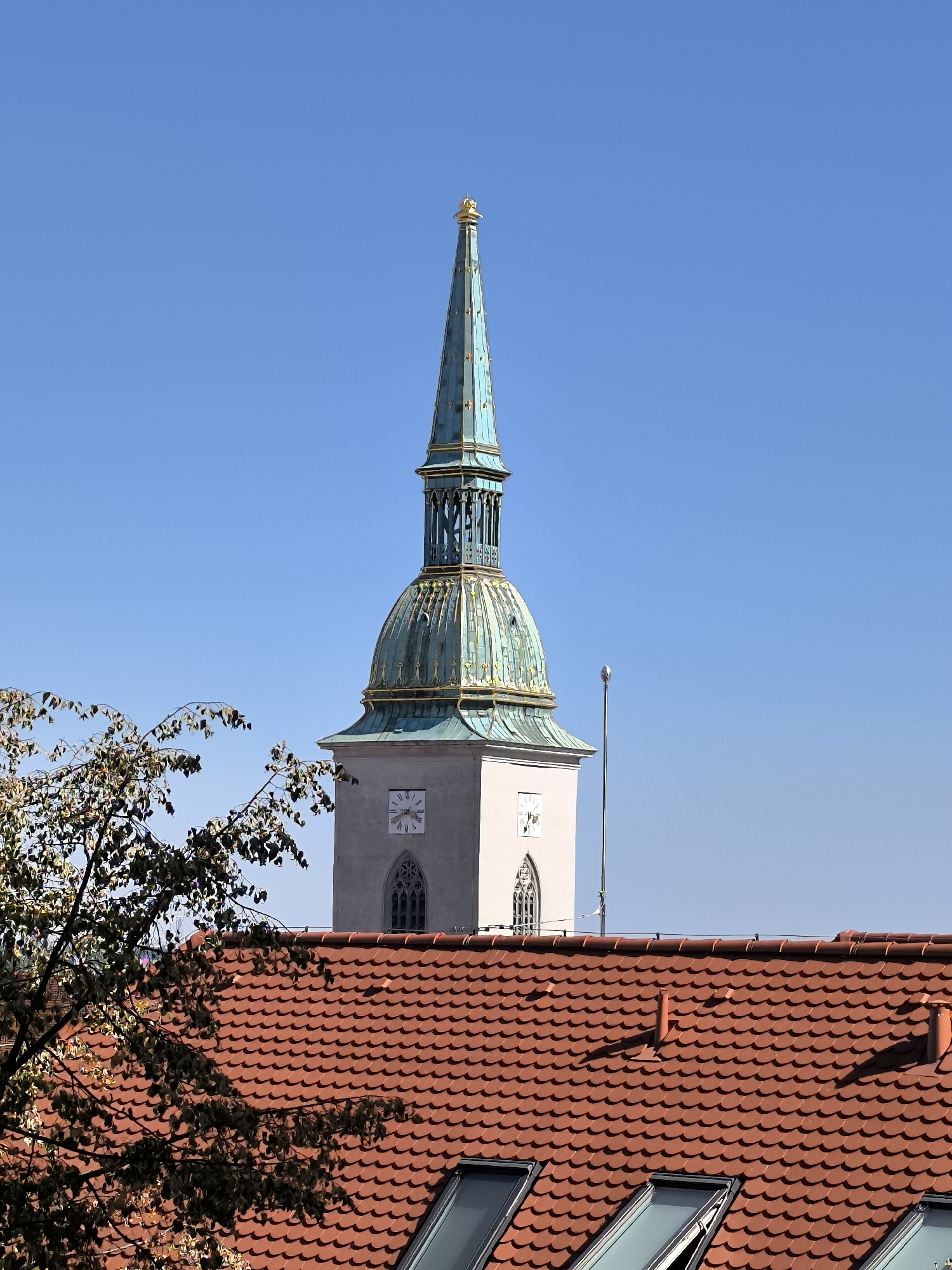 This is a medium shot, taken from an elevated perspective, of the St. Martin's Cathedral tower in Bratislava, Slovakia. The tower, a prominent landmark, is rendered in pale stone with a copper spire that has a greenish patina. The spire is topped with a golden finial. The lower part of the tower features a clock face and a Gothic-style window. In the foreground, a section of a red-tiled roof with several skylights is visible. To the left, the branches of a tree with sparse, autumnal foliage extend into the frame. The sky above is a clear, vibrant blue. The scene appears to be an outdoor view on a bright, sunny day.