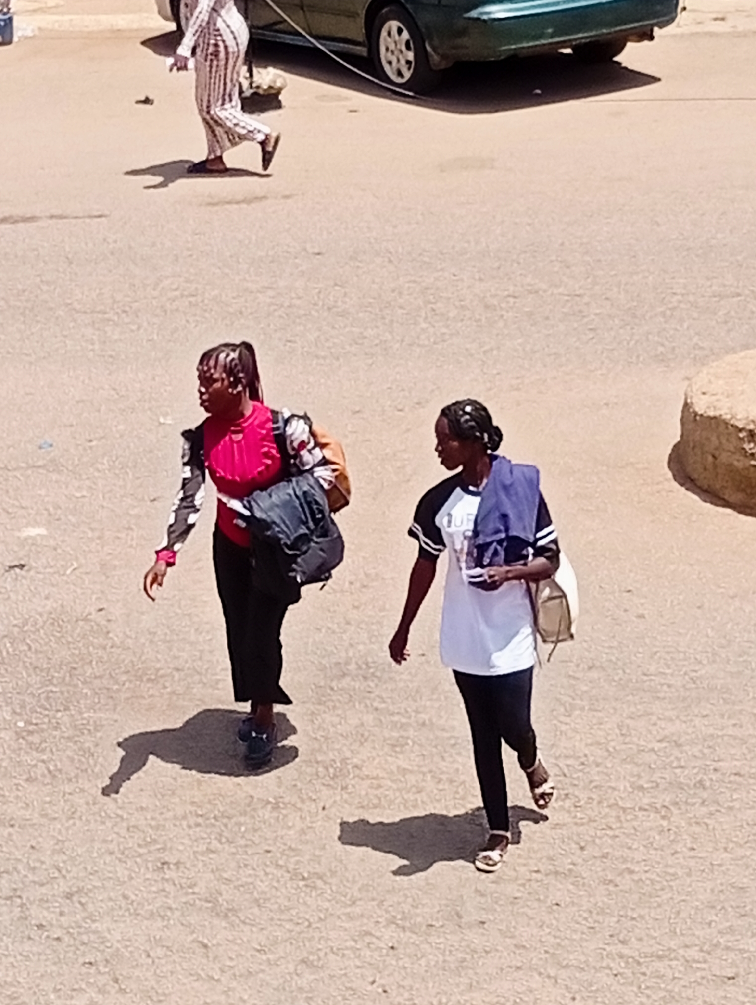 Two young women are walking on a paved road in Bukuru, Nigeria. The woman on the left is wearing a pink and black long-sleeved shirt and black pants. She is carrying a dark backpack and a brown bag over her left shoulder. The woman on the right is wearing a white t-shirt with black sleeves and black pants. She is carrying a light-colored bag over her right shoulder and a blue garment is draped over her left shoulder. A third person, partially visible in the background, is wearing a striped outfit. A green car is parked on the side of the road. The ground is paved and there is a large rock formation to the right of the frame. The sun is casting shadows on the ground.