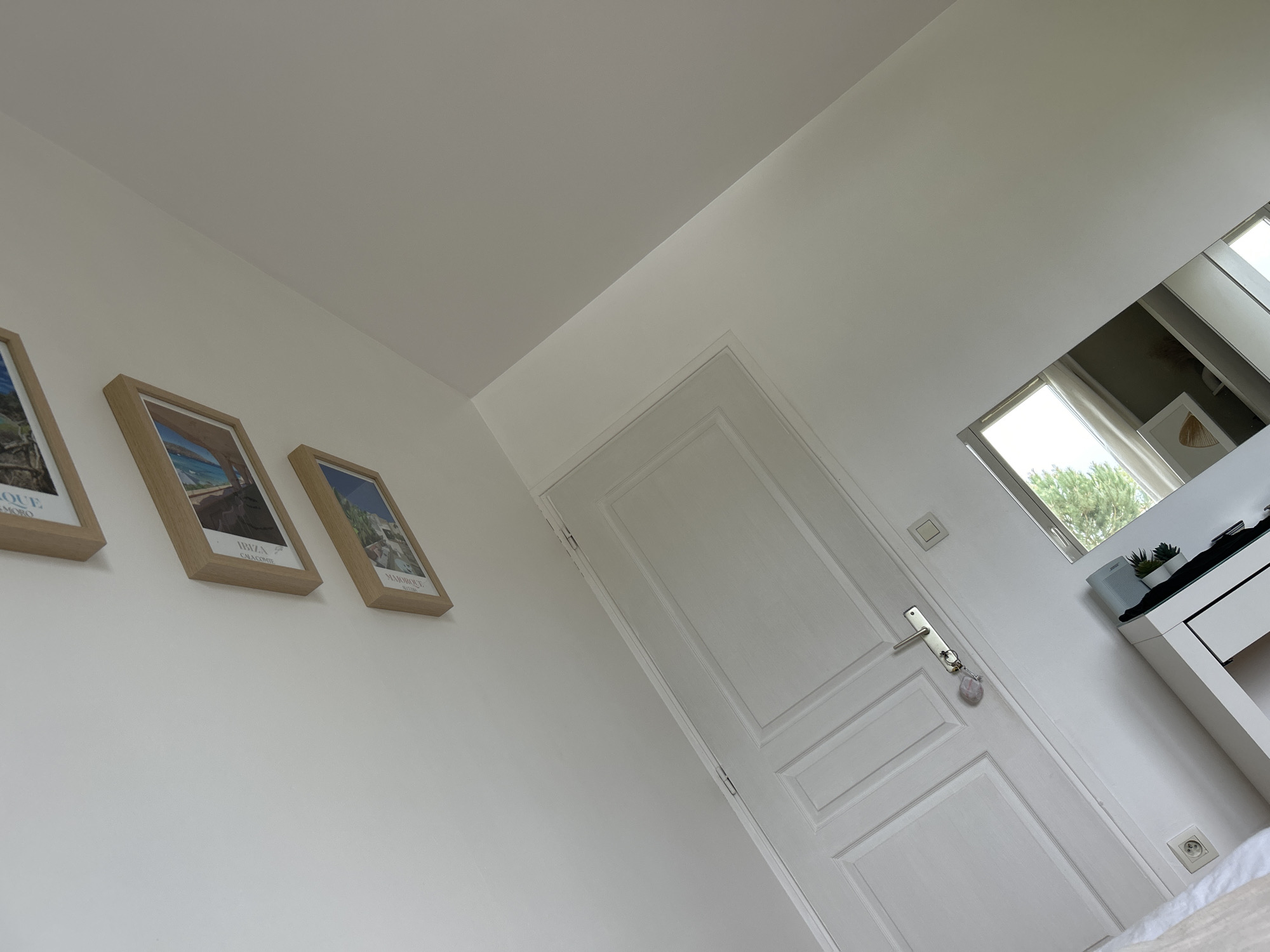 The image shows a section of an interior space. The walls are painted white, and there are three framed pictures hanging on the wall to the left. The frames are light wood, and the pictures depict landscapes or scenes. A white door with rectangular panels is centrally located and slightly angled. A door handle and what appears to be a keychain are attached. Adjacent to the door is a window with a view of greenery and a white desk. The reflection in the mirror suggests a modern interior design with muted tones. The scene is well-lit and appears to be from a residential setting. The location in Veauchette, France is not explicitly confirmed or evidenced from the content of the image.