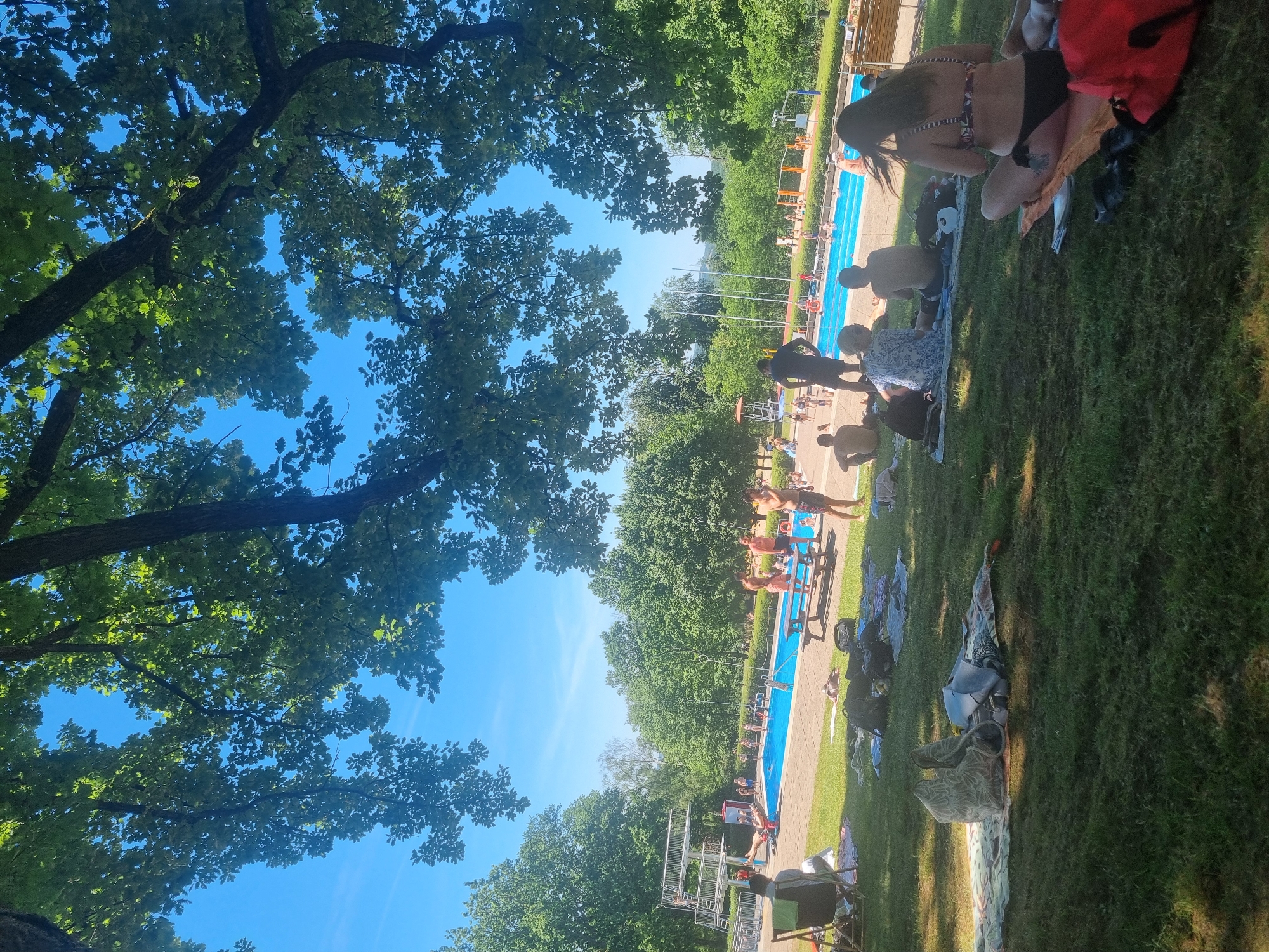 The image depicts a swimming pool scene in Saarbrücken, Germany. Patrons are seen sunbathing on the adjacent grassy area. Some individuals are in the water. The sky is clear and blue, partially obscured by trees in the foreground.