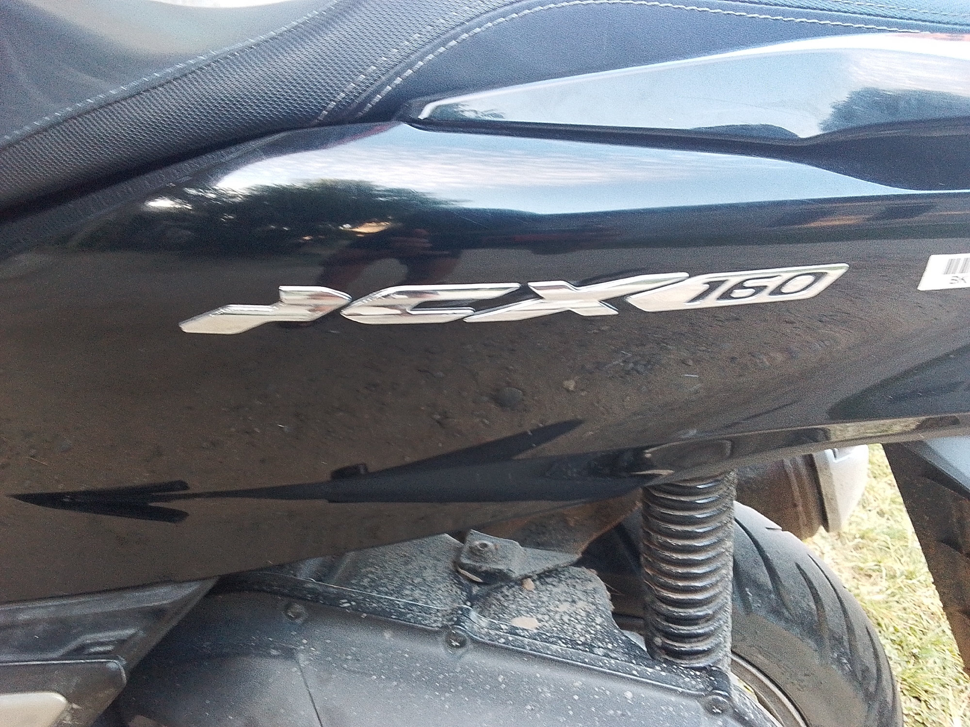 The image shows the rear section of a black Honda PCX 160 scooter. The "PCX 160" logo is visible in silver lettering on the body panel. Beneath the logo is the scooter's rear suspension, tire, and lower bodywork, showing signs of dirt and road grime. The image appears to be taken outdoors, and the reflection on the shiny scooter body panel suggests a bright, possibly overcast, day. The background reveals an area with vegetation and what appears to be a dirt or gravel surface. Based on the question context, the location is presumed to be Lhokseumawe, Indonesia.