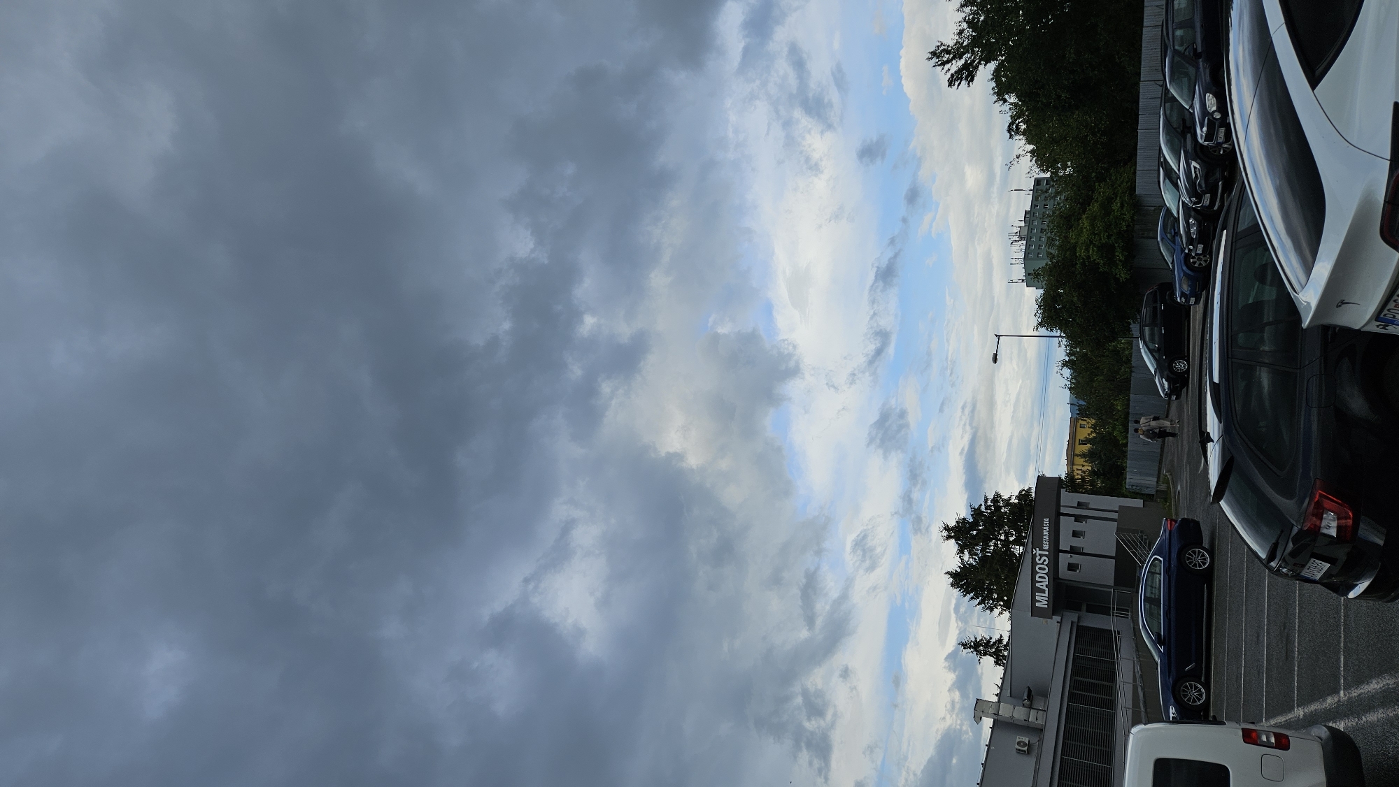 This is a vertical, slightly tilted, outdoor photograph of a parking lot with a building and trees in the background. The sky is dramatic, with a mix of dark, heavy storm clouds on the left and patches of bright blue sky with lighter clouds on the right.
In the parking lot, several cars are parked in rows. The cars visible include a white SUV or crossover, a black car, a dark blue sedan, and other darker vehicles further down the line. The white car on the right has a Tesla logo visible on its rear.
A modern-looking building with a sign that reads "MLADOST' REŠTAURÁCIA" is partially visible on the left. The building appears to have multiple levels and some windows. Trees line the edge of the parking lot and surround some of the buildings.
Two people are walking in the distance near the building, appearing small in the frame.
The overall impression is that of a parking area on an overcast day, possibly before or after a storm, given the contrast between the dark and bright parts of the sky. The lighting suggests it is likely late afternoon or early evening. The scene is set in an urban or suburban environment with a mix of commercial buildings and greenery.