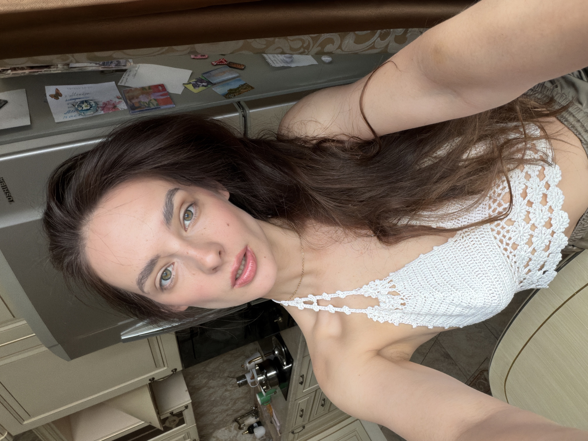 The image is a selfie taken in a kitchen. A young woman with long brown hair and green eyes is looking at the camera. She is wearing a white crochet top and has a gold necklace on. In the background, there are kitchen cabinets, a refrigerator with magnets on it, and a stove with pots and pans. The environment appears to be domestic. There is no information in the image to suggest a specific location in Ufa, Russia.