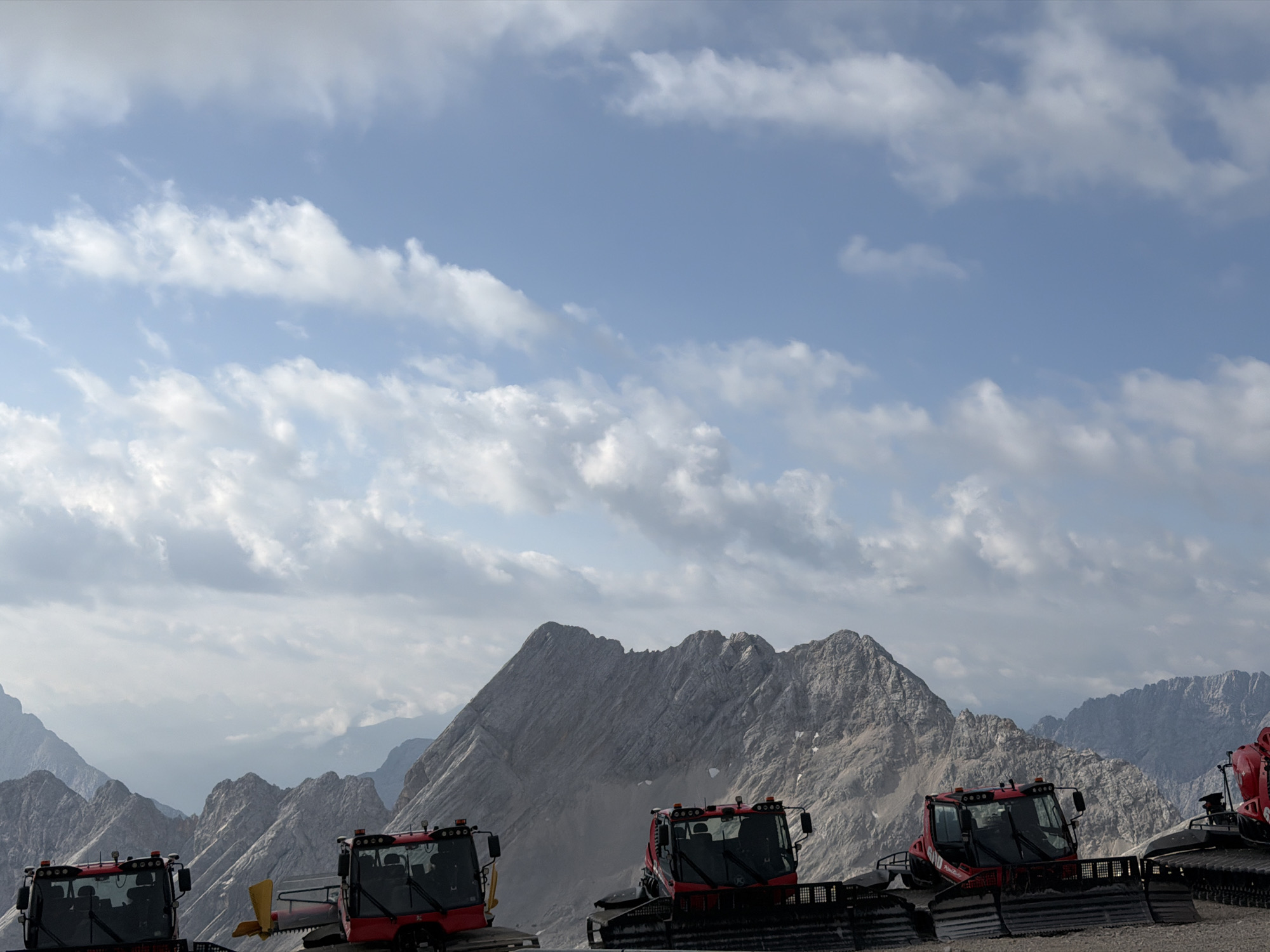 This image, taken in the vicinity of Garmisch-Partenkirchen, Germany, depicts a scenic mountain landscape under a partly cloudy sky. In the foreground, several red snow grooming machines are parked on a dirt or gravel surface. These machines, with their distinctive tracks and cabins, suggest a mountain resort or a preparation area for winter sports. The background is dominated by rugged, rocky mountain ranges, with a prominent, sharp peak in the center. The sky features a mix of blue sky and scattered white clouds. The overall impression is one of a high-altitude, natural environment, likely during the daytime. No people are visible in the frame.