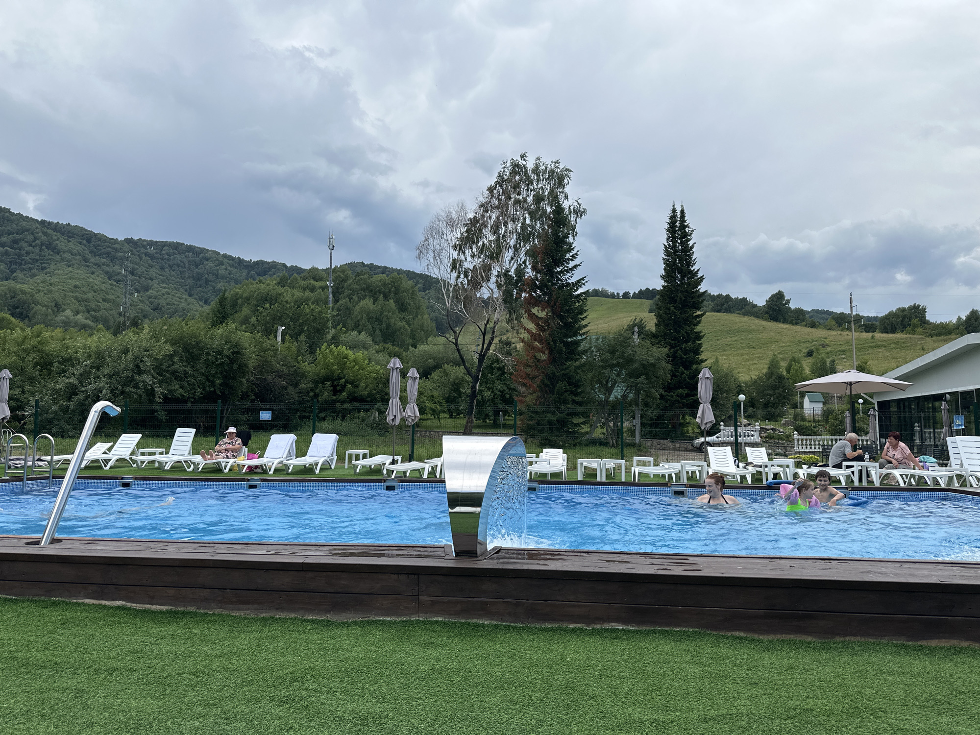 The image depicts a swimming pool area at a resort, possibly in Danilovka, Altai Krai, Russia, under a cloudy sky. The pool is filled with blue water and has a modern silver waterfall feature. Several people are visible: two children and an adult are in the pool, with the children wearing arm floaties. On lounge chairs around the pool, a woman wearing a hat is relaxing, and further back, a man and two women are seated. White lounge chairs, umbrellas, and tables are arranged on a wooden deck surrounding the pool, which transitions to a bright green artificial turf area in the foreground. In the background, a lush green forested hillside rises, with a few trees and some communication towers visible on the slopes. A white building with a sloped roof and outdoor seating area is partially visible on the right side of the image. The overall scene suggests a recreational setting with overcast weather.
