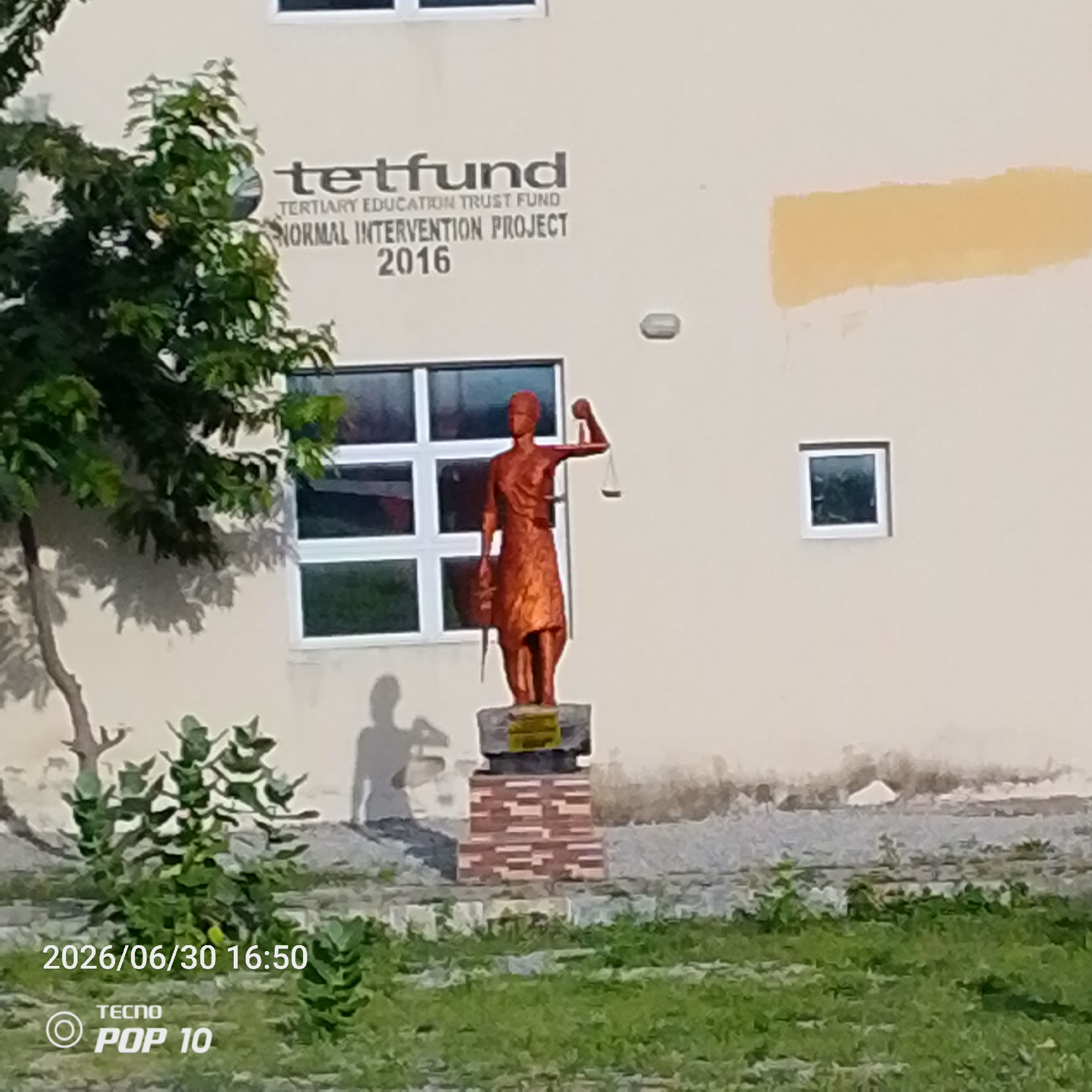 The image shows a statue of a woman holding scales, a symbol of justice, in front of a building. The statue is made of copper-colored material and stands on a brick pedestal. Behind the statue, there are large windows with white frames. To the right of the statue, a small square window is visible. The building has a light-colored facade with some text printed on it. The text reads "tetfund" followed by "TERTIARY EDUCATION TRUST FUND" and "NORMAL INTERVENTION PROJECT 2016". A large tree with green leaves is on the left side of the image, casting a shadow on the building. The foreground is filled with green grass and some plants. In the bottom left corner of the image, a timestamp "2026/06/30 16:50" and "TECNO POP 10" are visible. The scene appears to be outdoors during the day, with clear weather. The statue suggests the location might be related to a legal institution or an educational facility that values justice. The time stamp indicates a future date and time. The setting is likely Jalingo, Nigeria, as per the provided context.