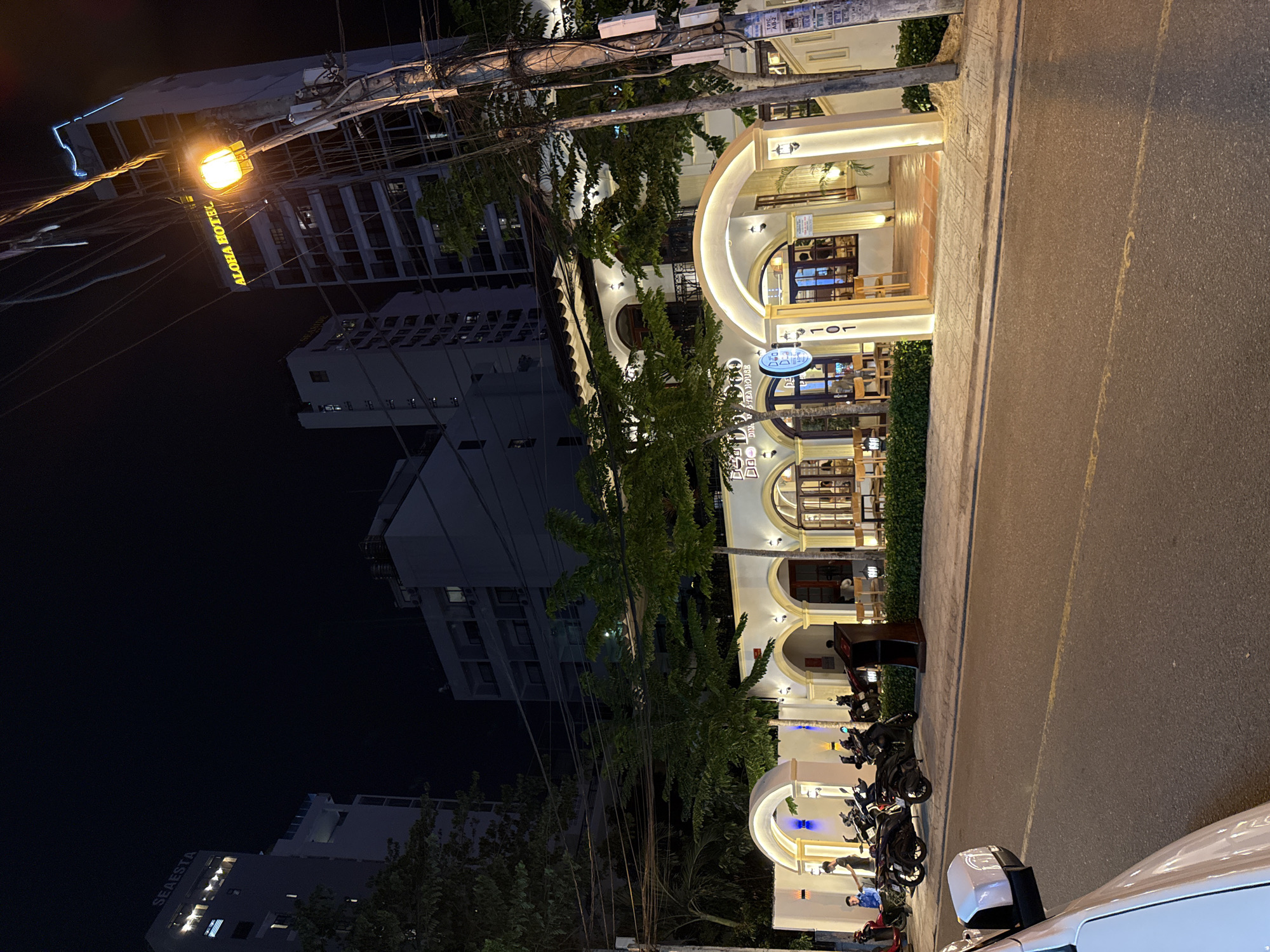 This image depicts a nighttime street scene in Nha Trang, Vietnam. The foreground shows a paved road with a white car partially visible on the left. In the mid-ground, a low-rise building with arched entrances and illuminated windows is prominently featured. Trees with lush green foliage are planted in front of this building. Several motorcycles are parked on the sidewalk. In the background, tall buildings, including the "SEAESTA" and "ALOHA HOTEL," are visible against the dark night sky. The scene is lit by streetlights and the interior lighting of the building.