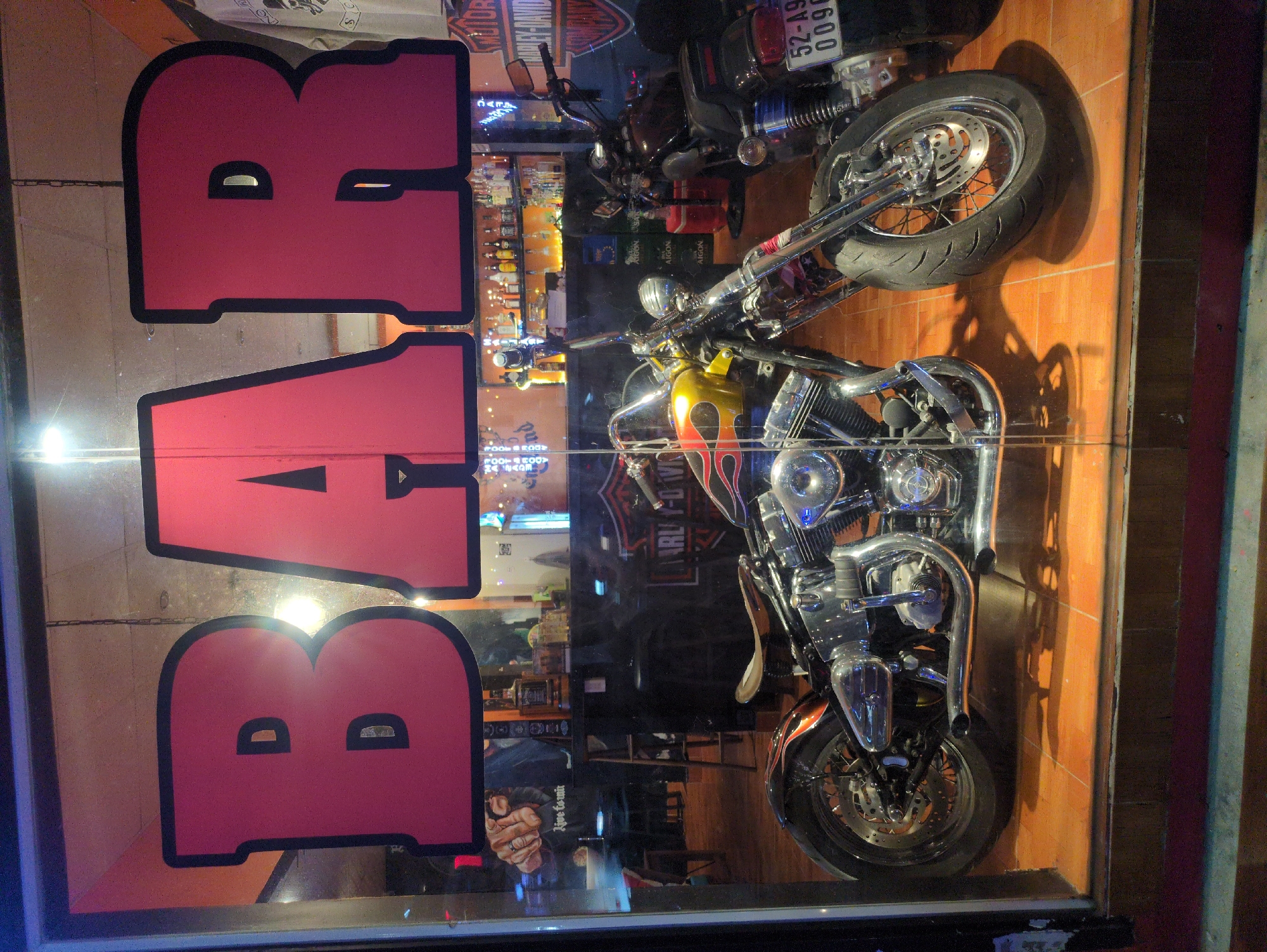 The image depicts a storefront window in Phú Quốc, Vietnam, with a prominent pink "BAR" sign divided into two parts. The reflection in the window reveals the interior of the bar, including a shelf full of liquor bottles and tables and chairs. In front of the window, two motorcycles are parked on the tiled floor, one with flames painted on the gas tank. Text on the motorcycles include “HARLEY-DAVIDSON” and a license plate number “52-A9 0096”. A hand reflected in the lower left portion of the window shows a person wearing a ring. A sign reading "Live tounir" is also partially visible inside the bar.