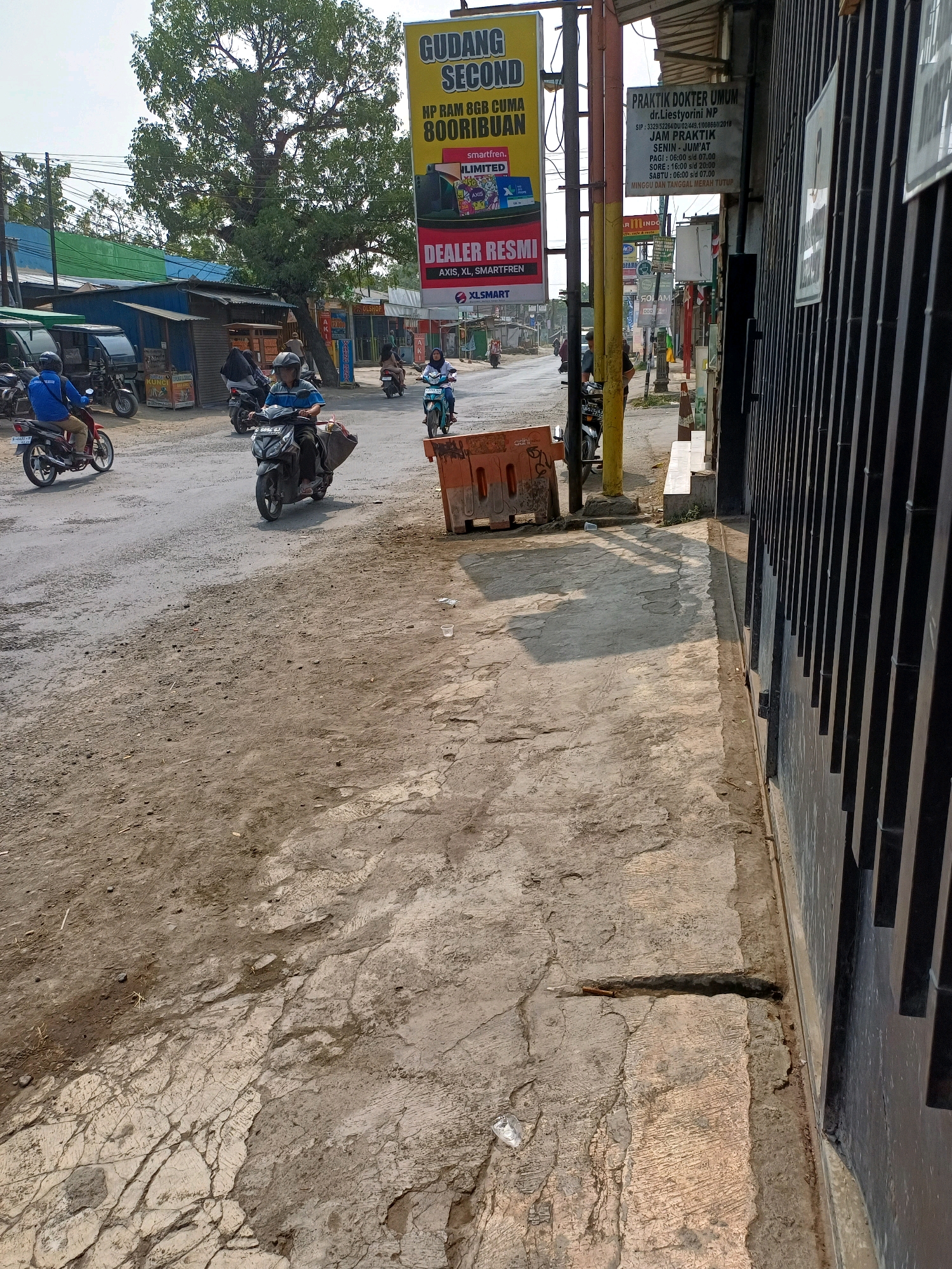 The image depicts a street scene in Kersana, Indonesia, characterized by a dusty, unpaved road with patches of cracked concrete. On the right side of the image, a dark, vertical barred gate lines the edge of a building. A yellow pole stands near the gate, supporting a sign for "PRAKTIK DOKTER UMUM dr. Liestyorini NP" which details the doctor's practice hours. To the left of the street, a large tree with dense foliage is prominent. Several shops are visible along the street, including one with a yellow banner advertising "GUDANG HP RAM 8GB CUMA 800RIBUAN". Motorbikes are a common mode of transport, with several people riding them on the road. One rider in blue is on the left, another in dark blue is in the center, and a rider in light blue is further down the street on the right. Orange barriers are placed on the side of the road. The environment appears to be a commercial or mixed-use area with shops and residences. The overall impression is a bustling, everyday street scene in a town in Indonesia.