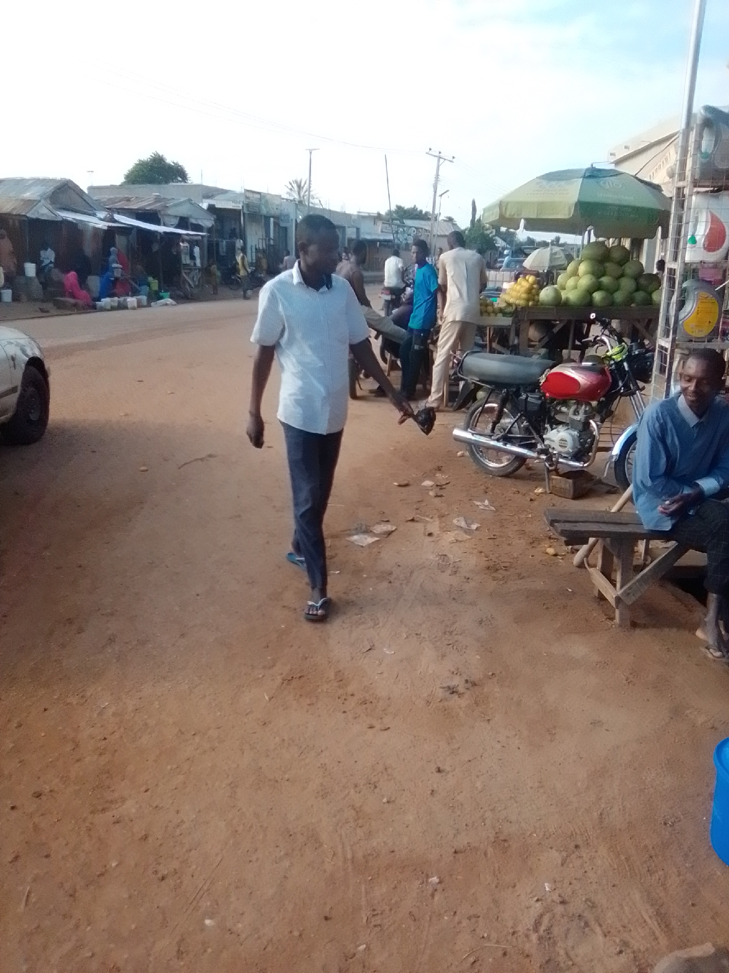 The image depicts a street scene in the city of Galadima, Nigeria, likely a market area. In the foreground, a man wearing a white short-sleeved shirt and dark trousers is walking away from the camera. To the right, another man is seated on a wooden bench, wearing a blue shirt and dark trousers, looking towards the left. Behind him, a red motorcycle is parked. In the background, there are several market stalls with corrugated metal roofs, and a large green and yellow umbrella shading a fruit stand displaying numerous green fruits, likely melons, and yellow fruits. Several people are visible in the background, some sitting and some standing around the stalls. The ground is unpaved and appears to be made of dirt or sand. A white car is partially visible on the left side of the image. The sky is overcast.