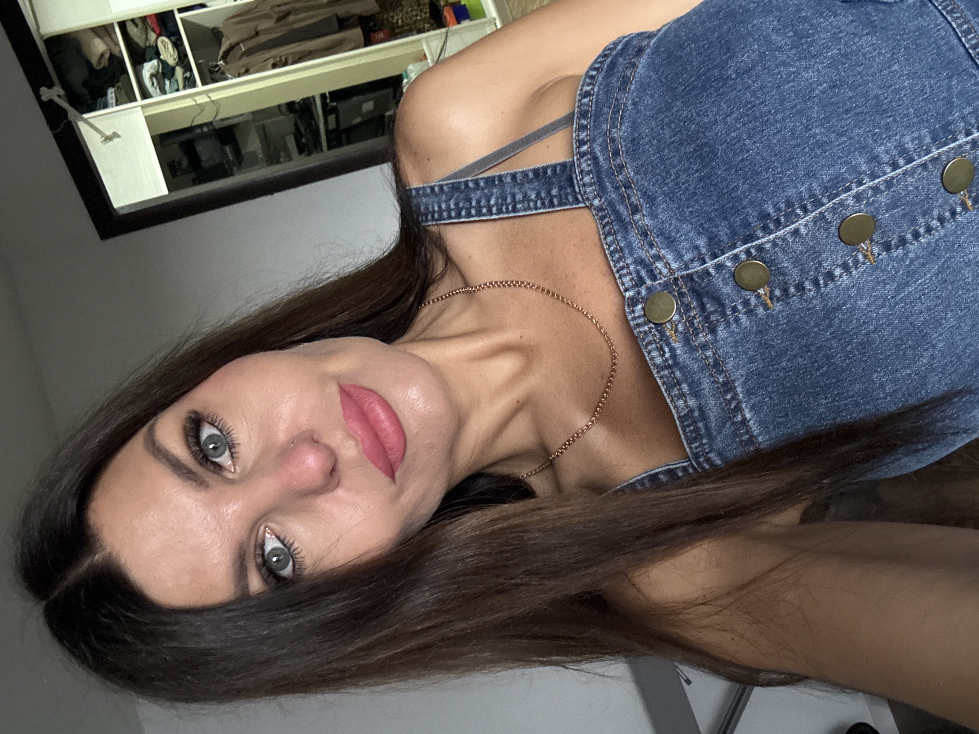 A medium shot shows a woman with long, dark brown hair and blue eyes looking at the camera. She is wearing a denim dress with gold buttons and a thin gold chain necklace. Her lips are painted a matte pink. In the background, a closet is visible, filled with folded clothes and hanging garments. The room appears to be a bedroom. The specific location in Chaikovsky, Russia is not discernible from the image.