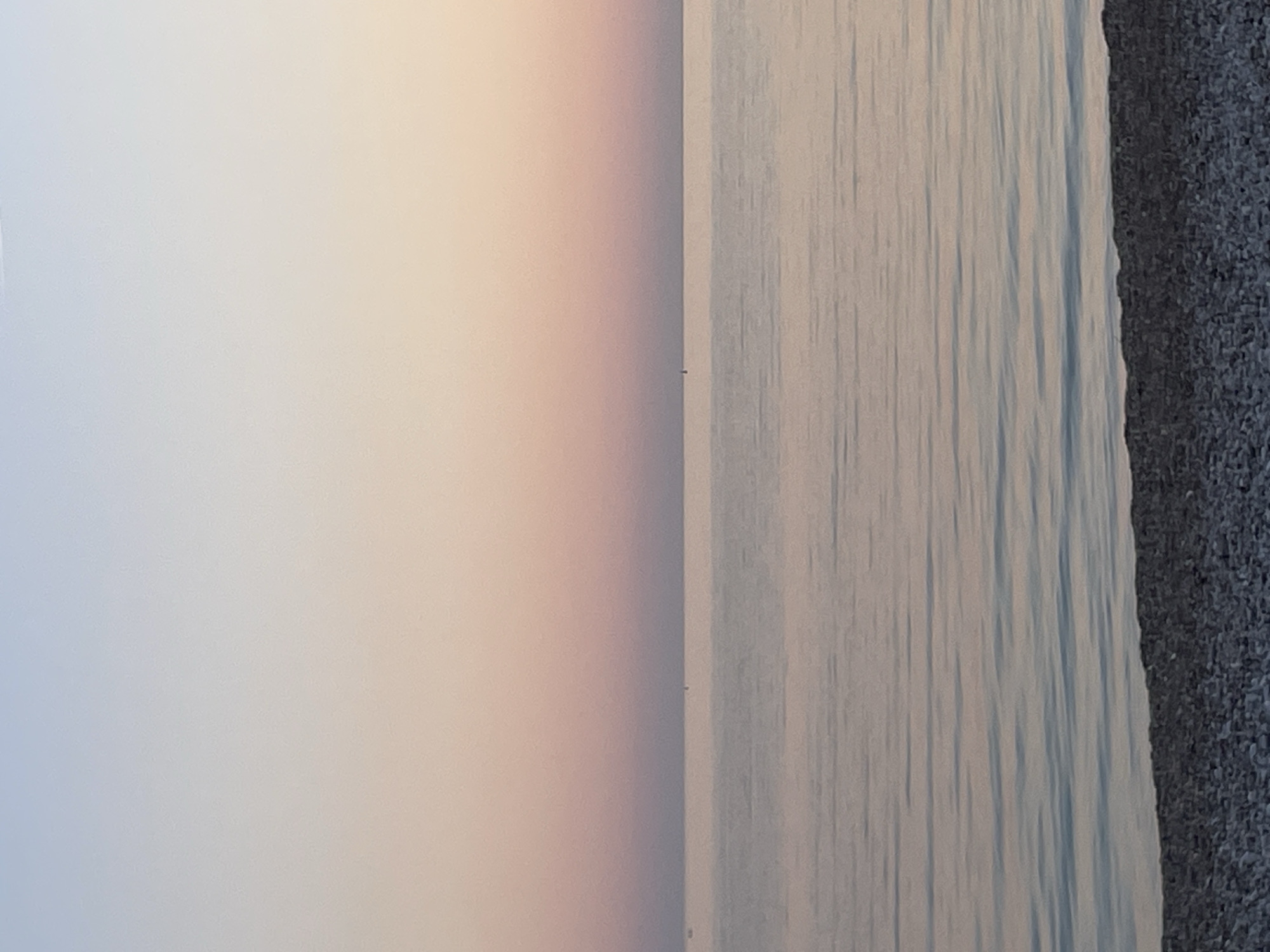 The image depicts a horizontal view of the sky, the sea, and the beach at Budva, Montenegro. The sky transitions from a pale blue to a soft pink-orange hue. The sea exhibits subtle ripples and reflections, with shades of light blue and gray. The beach is covered with fine, dark pebbles. No people or other objects are visible.