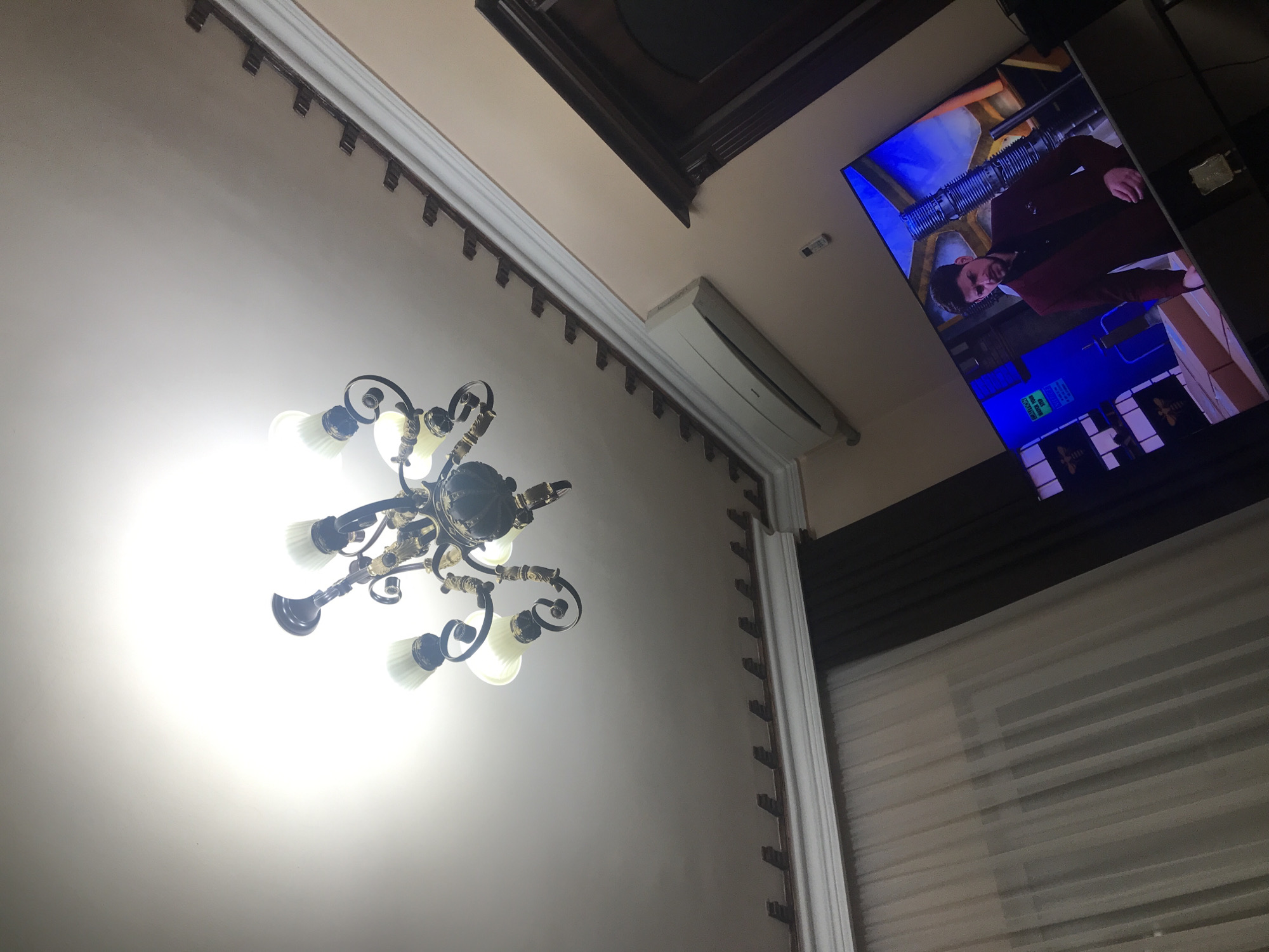 The image depicts an interior view, likely in Yerevan, Armenia, featuring a ceiling-mounted chandelier with multiple illuminated bulbs as the central focus. Adjacent to the chandelier, there's a wall-mounted air conditioning unit. A television screen displays a scene with a man wearing a dark red coat. The room also has a window covered with blinds. The ceiling is off-white with decorative molding.