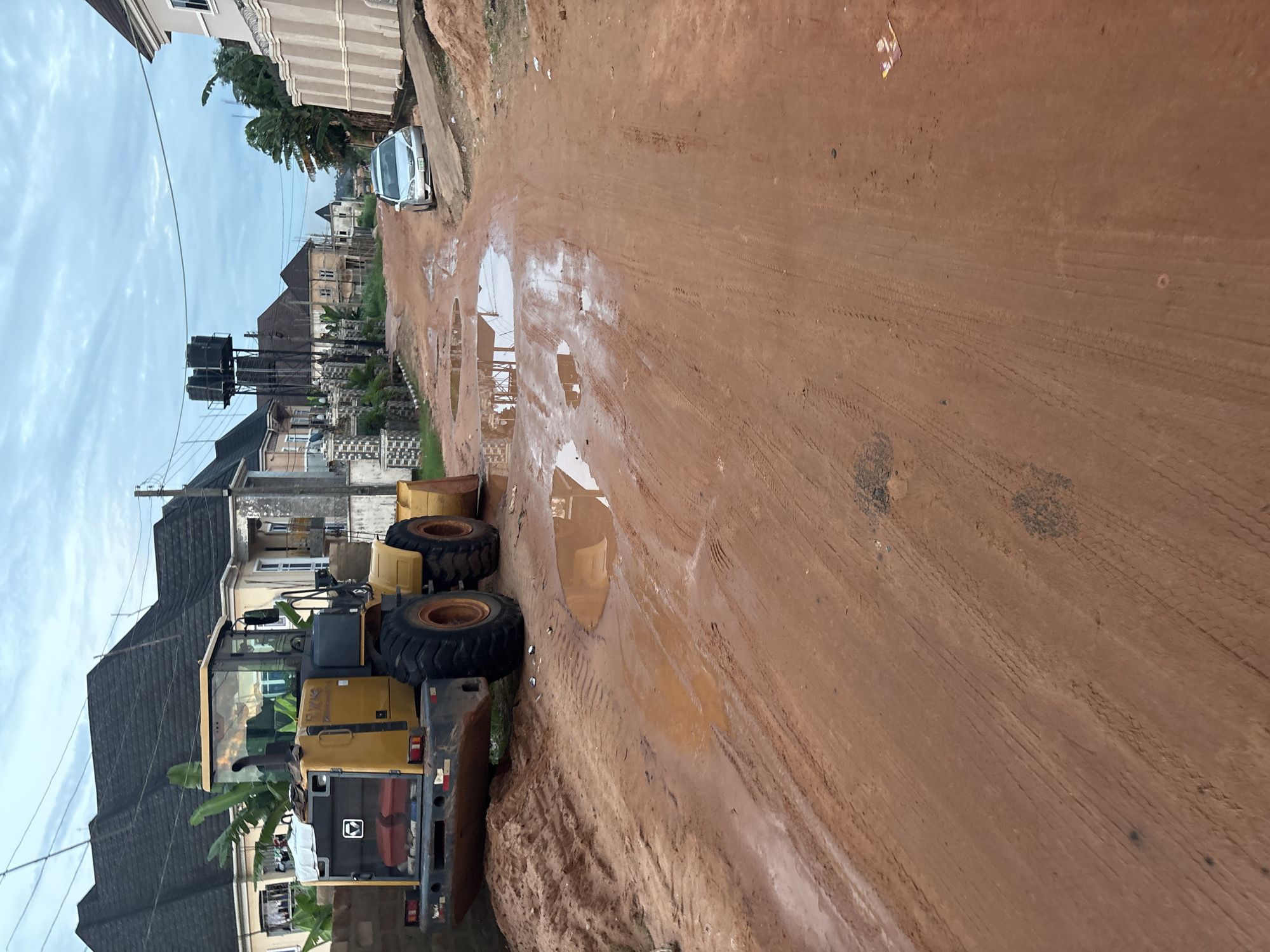 In Obosi, Nigeria, a yellow wheel loader is parked on a muddy, unpaved road. The road is pocked with numerous puddles, reflecting the overcast sky and surrounding buildings. To the left, houses with dark-tiled roofs are visible, some with visible construction materials. A silver sedan is parked further down the road on the right, next to a low wall. The environment appears to be a residential area undergoing some form of construction or maintenance, evidenced by the presence of heavy machinery and the state of the road. There are no people visible in the scene.