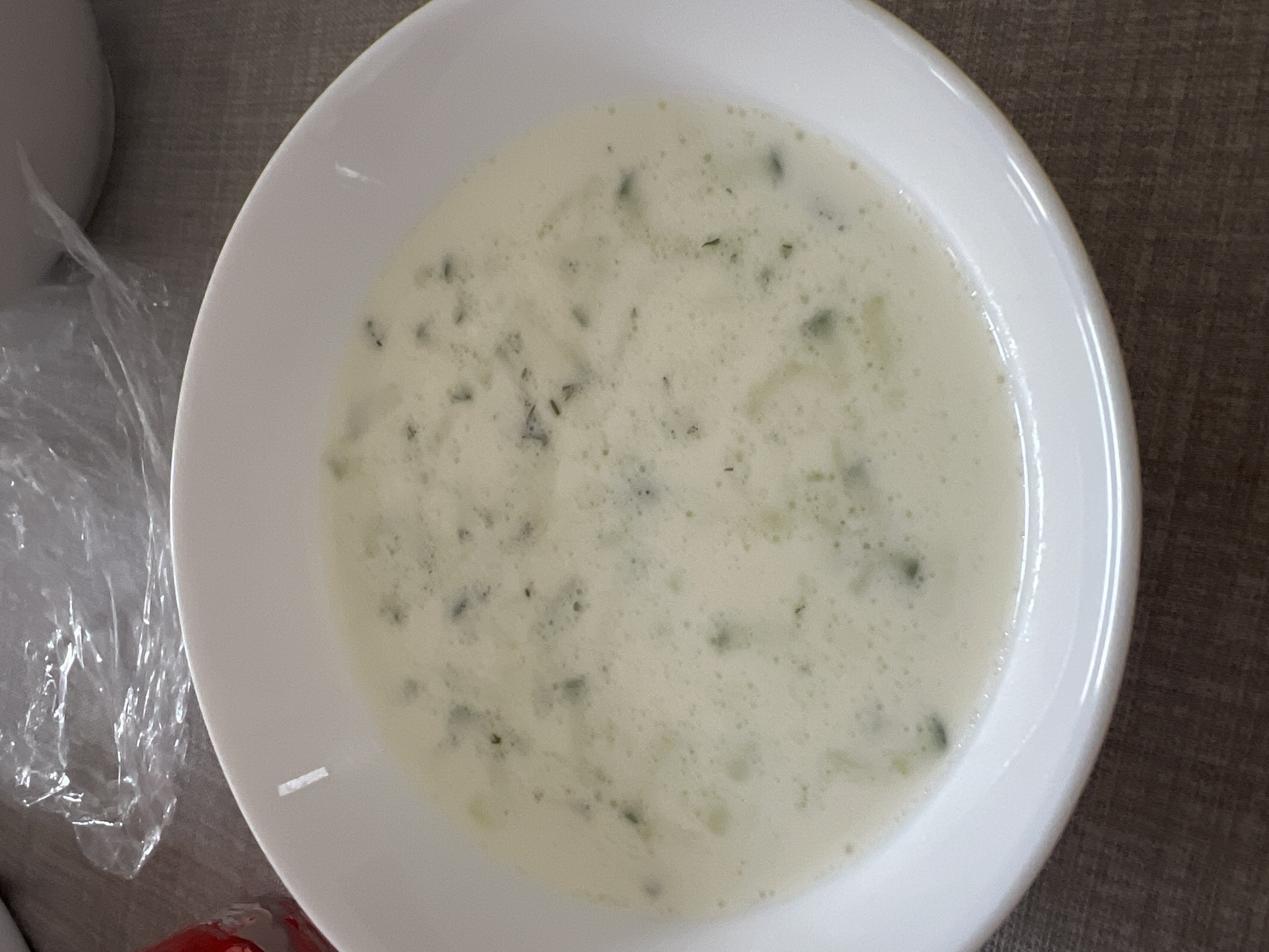 A close-up shot of a white bowl filled with a creamy soup, likely Tzatziki or a similar yogurt-based soup with herbs and potentially cucumber. The bowl is on a brown surface, and there is some plastic wrap or packaging partially visible on the left side of the image.