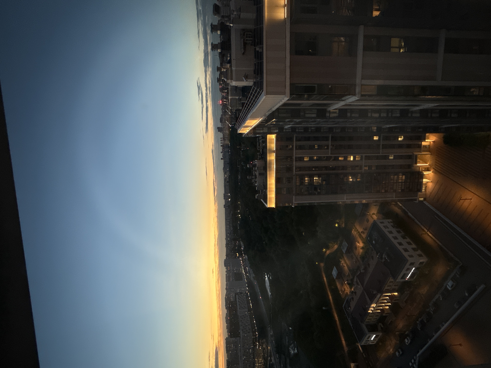The image captures a twilight view over a cityscape, likely Dolgoprudny, Russia, from a high vantage point. In the foreground, two modern residential buildings stand tall, their upper floors illuminated with warm, decorative lighting. The building on the right is closer and shows a side view, revealing multiple windows and structural elements. The building to its left is seen from a slightly more elevated perspective, showcasing its illuminated facade and the surrounding outdoor lighting.
Below the residential buildings, a darker area of trees suggests a park or green space. A road with visible car lights indicates traffic flow, hinting at the urban environment. In the mid-ground, a complex of older, lighter-colored buildings with columns is illuminated by outdoor lights, casting warm glows on its facade and the surrounding parking lot where several cars are parked.
The sky transitions from a deep blue at the top to a soft yellow and orange hue near the horizon, typical of sunset or sunrise. Faint cloud formations are visible, and a subtle, arc-shaped atmospheric phenomenon, possibly a faint rainbow or optical effect, is present in the upper sky. The overall scene depicts a residential area within a city during the transitional light of dusk.