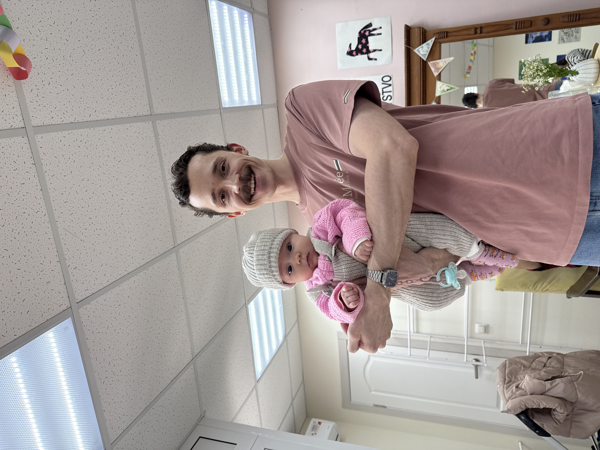 A medium shot features a man holding a baby. The man has dark hair, a mustache, and is wearing a mauve-colored t-shirt and jeans. The baby is wearing a grey knit hat and a pink cardigan. The man is holding the baby in his arms, facing the camera. A baby pacifier is visible. The background includes a ceiling with fluorescent lights, a wall painted a light pink color, a mirror hanging on the wall, and a door. The location appears to be inside a building in Chaikovsky, Russia, based on the Cyrillic script visible on the wall.