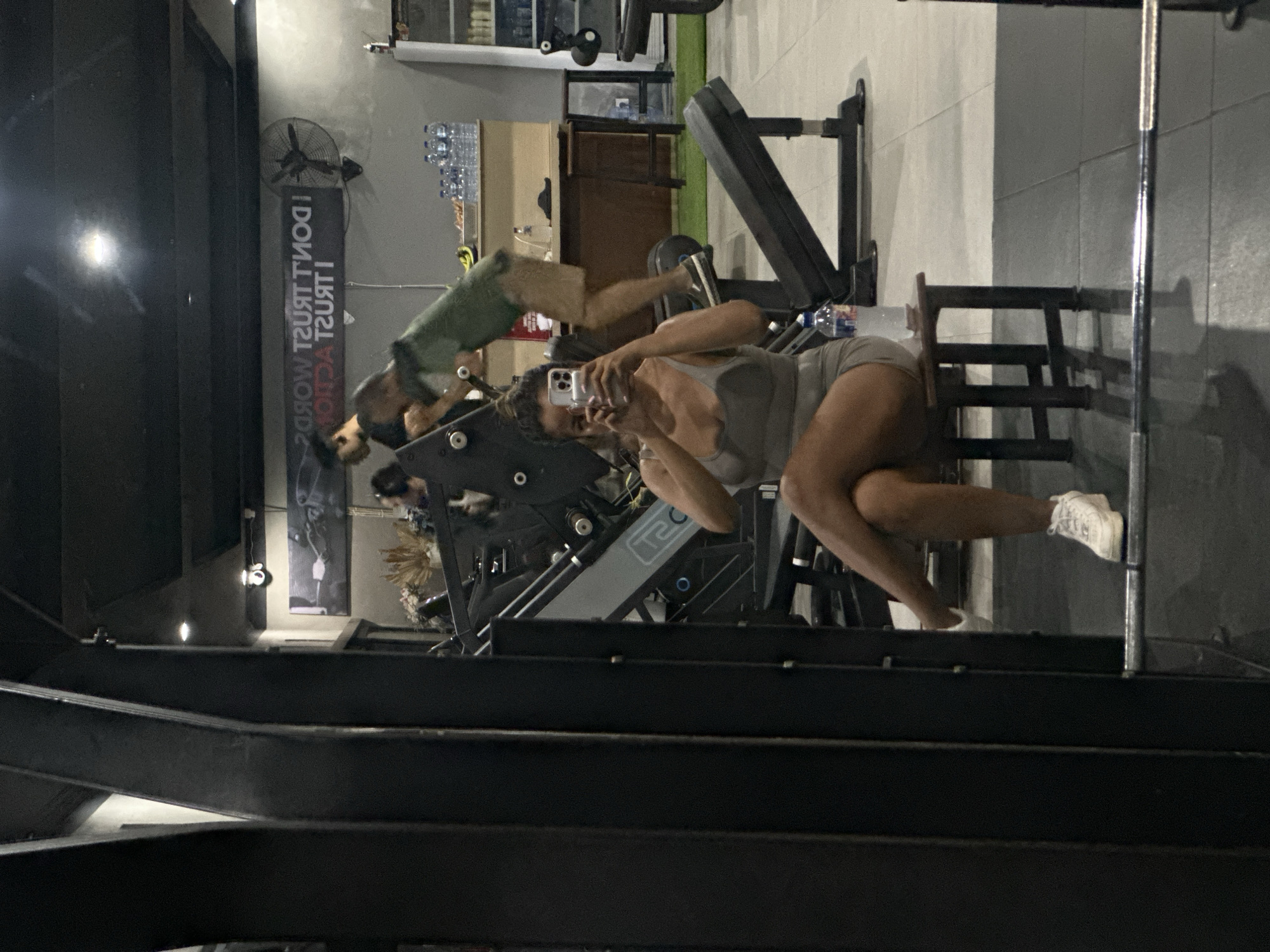 A young woman with dark hair pulled back is sitting on a small stool in a gym, taking a selfie with her phone. She is wearing a grey sports bra and grey shorts. Her legs are crossed, and she has white sneakers on her feet. A water bottle is on the stool next to her. In the background, through a large mirror, other gym-goers are visible. One man is using a leg press machine, and another man is in the background, partially obscured. A sign with text is visible on the wall. The gym appears to be in Sukawati, Indonesia, based on the context provided. The environment includes gym equipment, a tiled floor, and a dark ceiling.