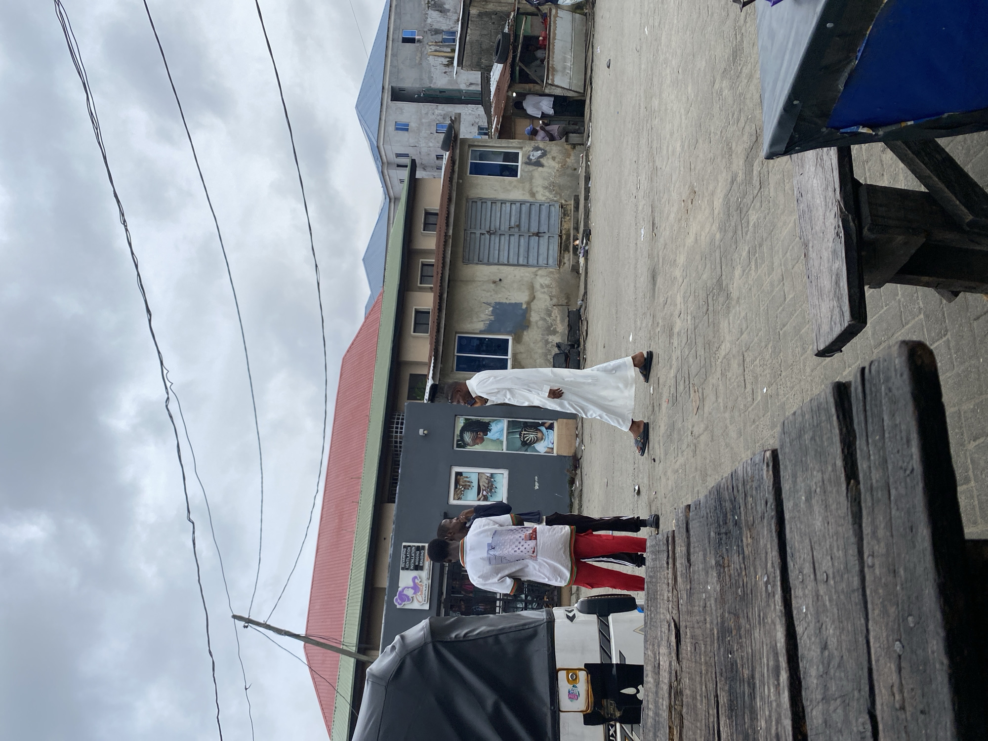 A cloudy day scene captures an outdoor street view with several people and buildings. In the foreground, a weathered wooden table and bench are visible, suggesting an outdoor seating area. Further back, a black trishaw is parked on the left. Three young men stand near the trishaw, two in casual wear and one in a white robe. To the right, a man in a white robe walks away from the camera, speaking on his phone. Behind them, a row of buildings with various architectural styles lines the street. Some structures appear older and less maintained, with weathered walls and corrugated iron roofs, while others are more modern multi-story buildings. The ground is paved with interlocking stones. The overall impression is a busy urban environment with a mix of commercial and residential structures under an overcast sky.
