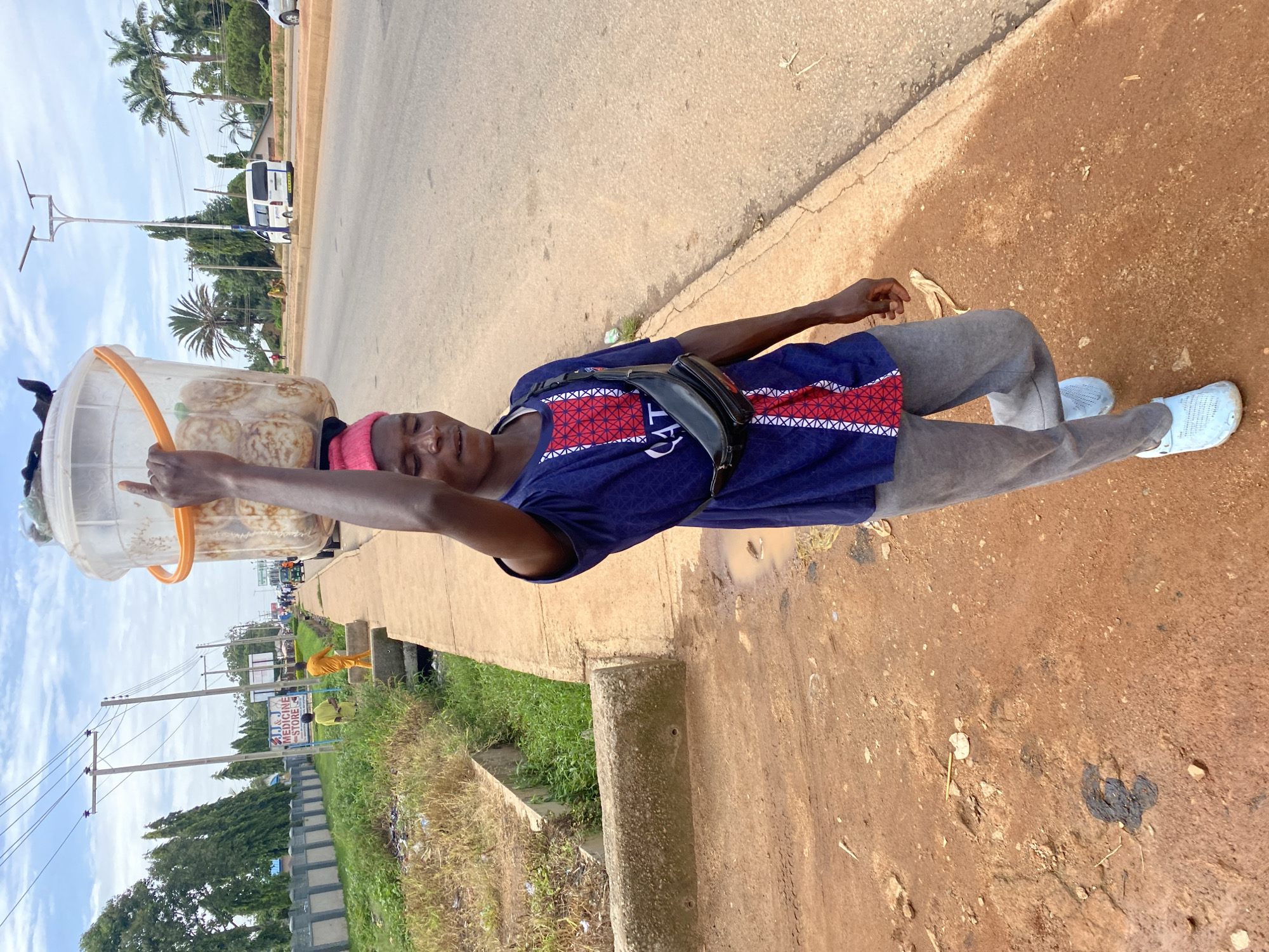 A person is standing by the side of a road, with their right arm outstretched and holding a large plastic bucket. The bucket appears to contain food items, possibly round pastries. The person is wearing a blue t-shirt with red and white trim, grey pants, and light blue shoes. They have a pink head covering and a black fanny pack across their chest. The person's eyes are closed, suggesting they might be resting or sleeping while standing.
The scene appears to be outdoors, with a paved road to the left and a dirt shoulder to the right. There are palm trees and other greenery in the background, as well as some buildings and power lines. A white van is visible on the road, and several other people are present in the distance, some wearing yellow clothing. The ground is a reddish-brown dirt, with some scattered debris. A banana peel is visible on the ground near the person's feet. The sky is cloudy.
The overall impression is of a street vendor taking a break during the day in a warm climate. The person's posture and closed eyes suggest fatigue or a moment of repose while still tending to their goods. There is a sign in the background that reads "J & J MEDICINE STORE." The lighting suggests it is daytime.