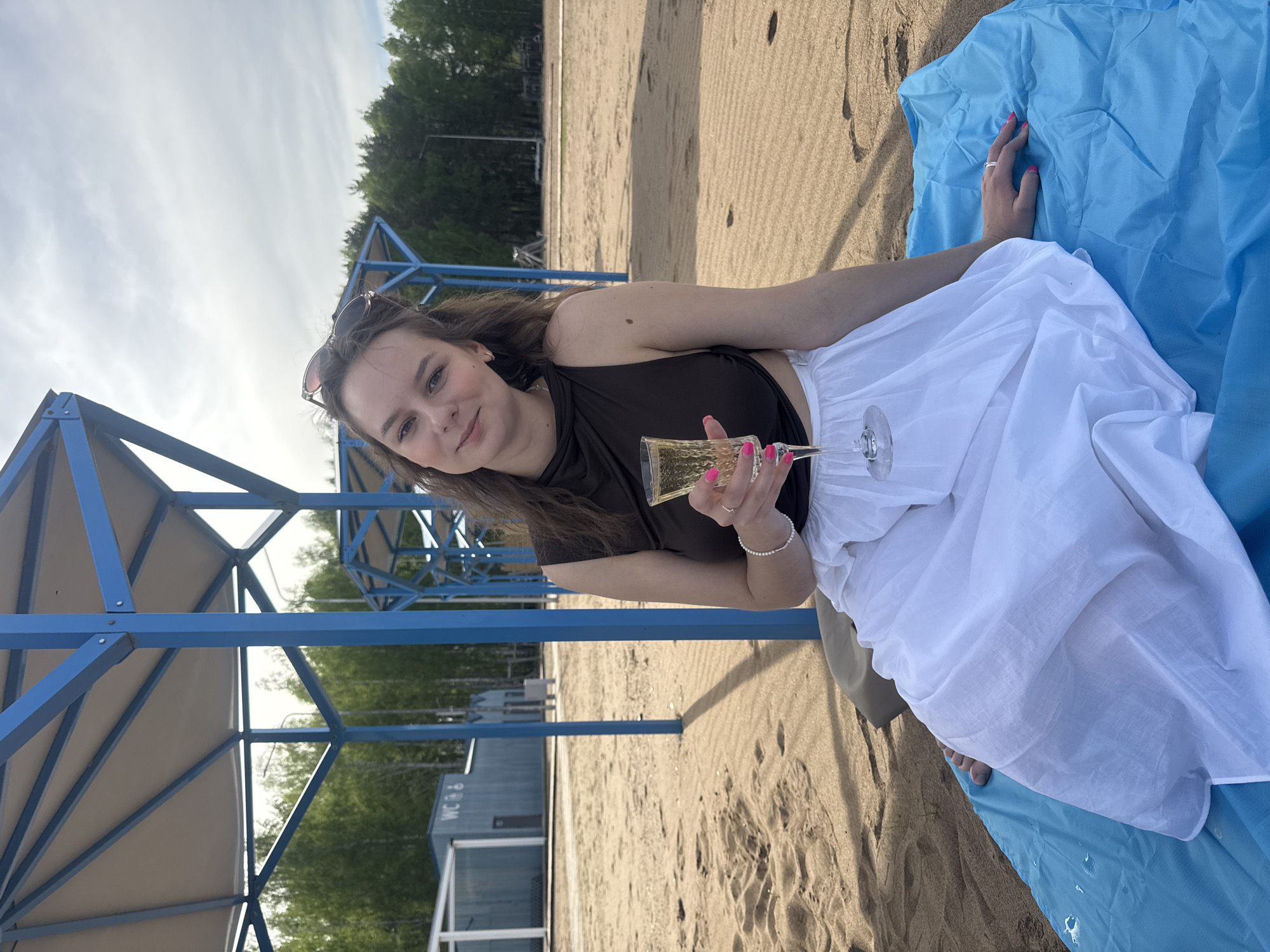 A young woman is sitting on a blue blanket on a sandy beach, holding a champagne flute in her right hand. She is wearing a dark-colored top and a white skirt. Sunglasses are perched on her head. The background includes beach umbrellas with blue frames and a building in the distance. The photo was taken in Chaikovsky, Russia.