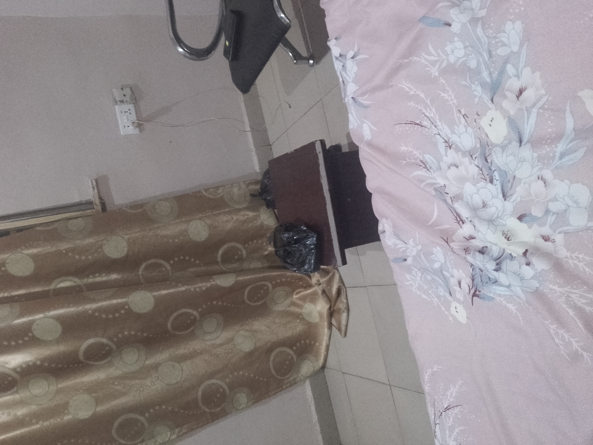 The image depicts a dimly lit bedroom interior. A light pink duvet with a floral pattern covers a bed on the right side of the frame. On the left, golden curtains with a circular pattern are drawn. A dark brown wooden nightstand is positioned between the bed and the curtains, with a black garbage bag placed on top. A white electrical outlet with a wire plugged into it is visible on the wall above the curtains. The floor appears to be tiled. The overall scene is cluttered and suggests a lived-in space.