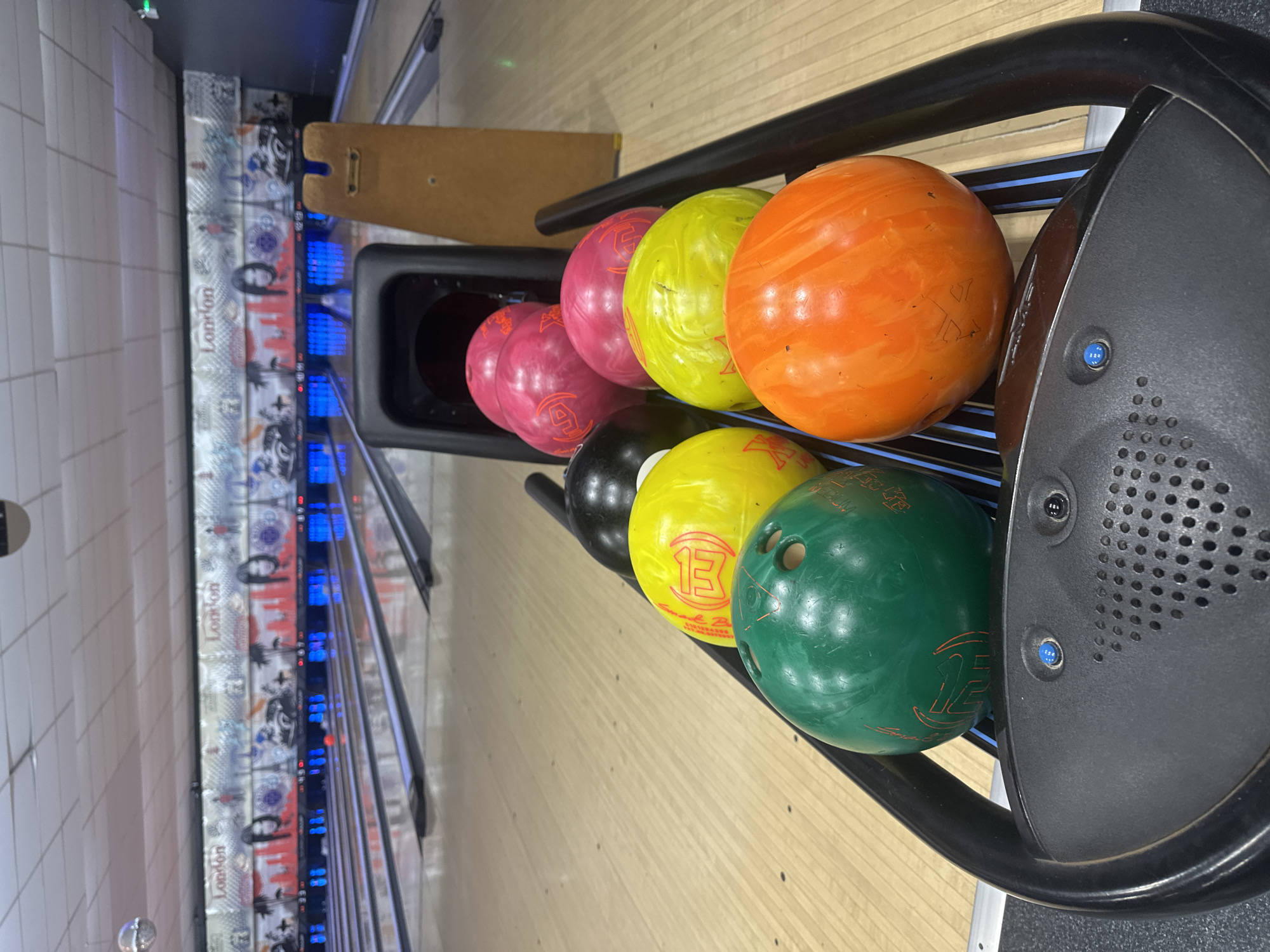 The image shows a bowling alley with multiple bowling balls lined up on a ball return rack. The bowling balls are of various colors, including green, yellow, pink, black, and orange. In the background, the bowling lanes are visible, with pins at the end of some lanes. The walls are decorated with murals depicting the city of London. There are no people visible in the image. The bowling alley appears to be indoors, with fluorescent lighting overhead. The overall scene suggests a recreational setting.