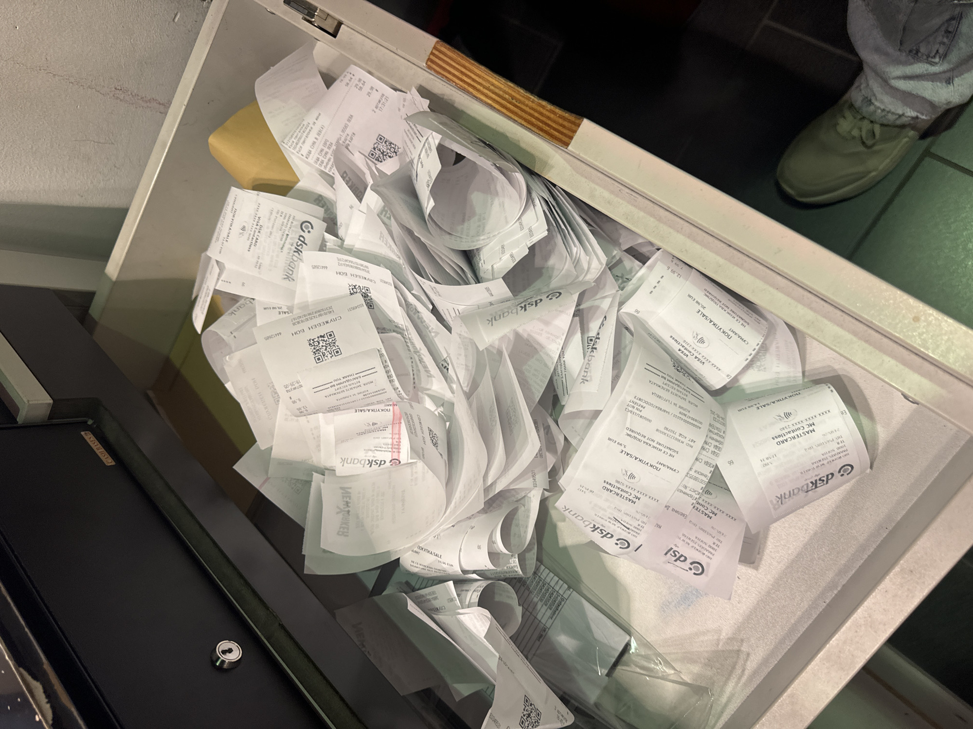 A cash register drawer is open, overflowing with numerous rolled and crumpled receipts, some bearing the logo "dskbank". The receipts are primarily white, with some having pink accents. A portion of the cash register is visible, including a lock on the drawer's face. The edge of a countertop is visible, with a foot wearing a green shoe standing beside the drawer. The location is unknown.