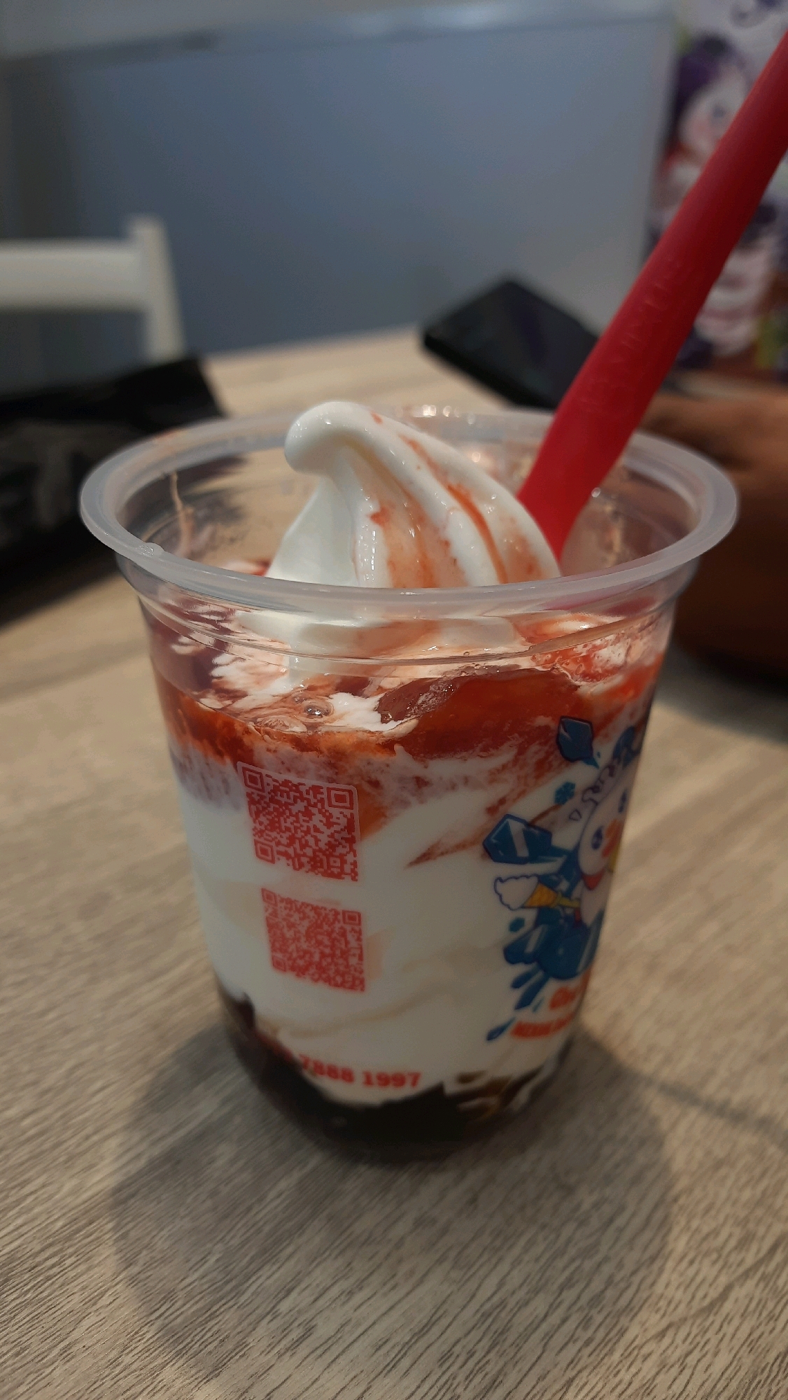 In Sidoagung, Indonesia, a close-up shot captures a clear plastic cup filled with soft-serve ice cream and strawberry sauce. A red spoon or straw is inserted into the ice cream, and a red QR code is visible on the side of the cup. The cup is placed on a wooden table. In the background, a blurred figure and a portion of a mobile phone can be seen.