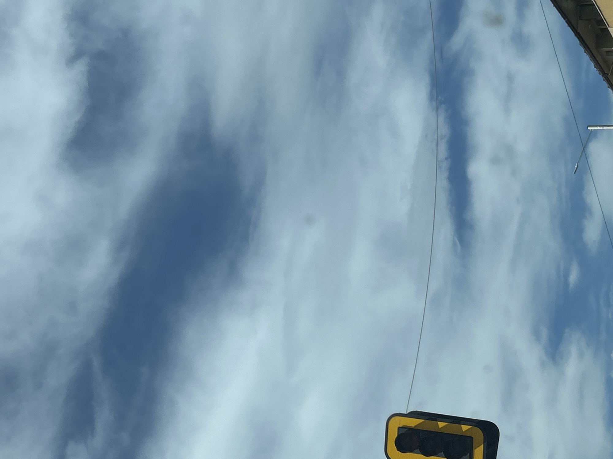 The image depicts a view upwards towards a cloudy sky in Sofia, Bulgaria. A traffic light is visible in the lower right corner. A portion of a building roof is seen in the upper right. Utility wires extend across the sky.