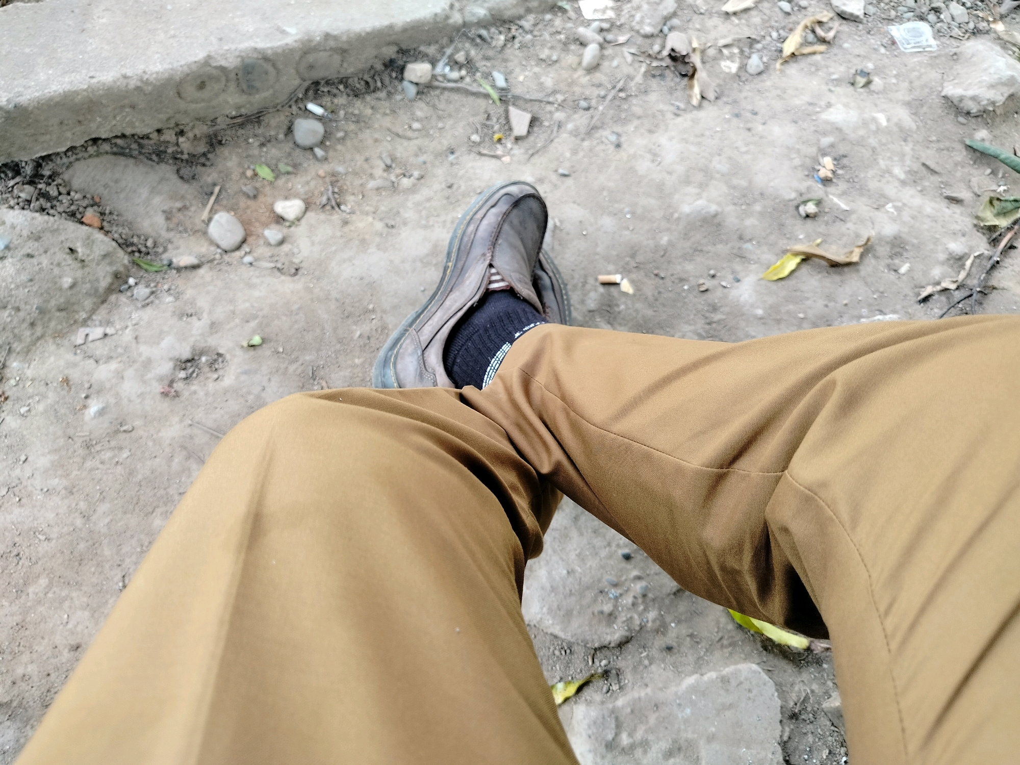 A first-person perspective shot shows a person sitting on a concrete ledge in an outdoor, possibly urban, environment. The person is wearing brown trousers and a dark sock. One brown leather shoe is visible, with the sock peeking out. The ground around the person is a mixture of dirt, small stones, and debris, including some dry leaves and what appears to be cigarette butts. In the background, more concrete and gravel can be seen, with some plant life. The lighting suggests it is daytime. The location is identified as Purwodadi, Indonesia.