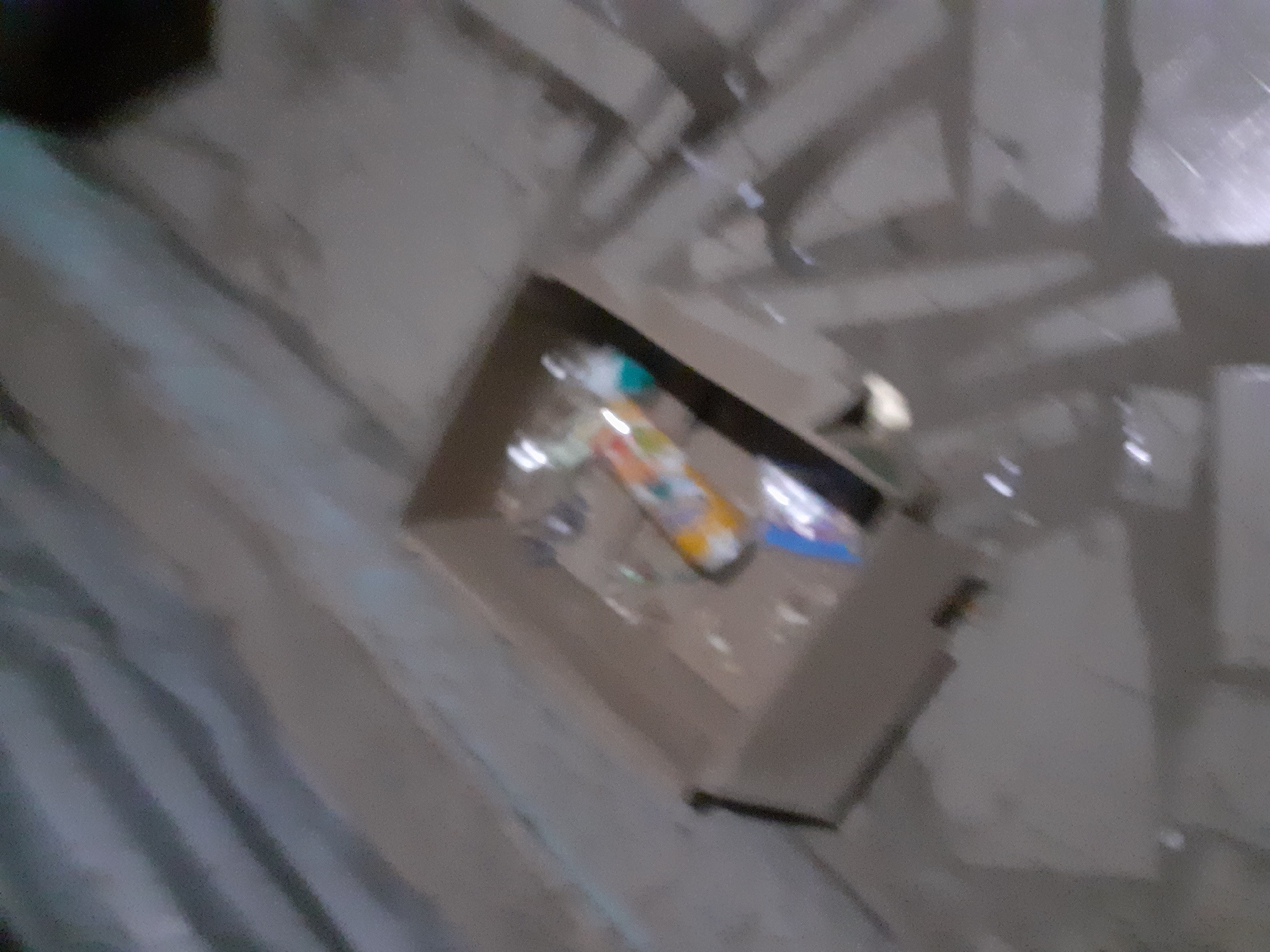 The image shows a close-up, blurry view of a cardboard box containing several objects, including what appear to be tubes or small containers with colorful labels. One object, possibly a tube of toothpaste or a similar personal care item, has a white base and a colored label with orange and yellow accents. Other items in the box are less distinct but suggest the presence of multiple small products. The box is placed on a light-colored, textured surface, possibly a floor, which is reflecting light. The background is also blurry and appears to be a continuation of the floor surface with some subtle patterns. The scene is indoors, and given the angle and lighting, it's difficult to determine the specific room or context. The overall impression is of a disorganized collection of items within a box. No individuals are visible in the image.
