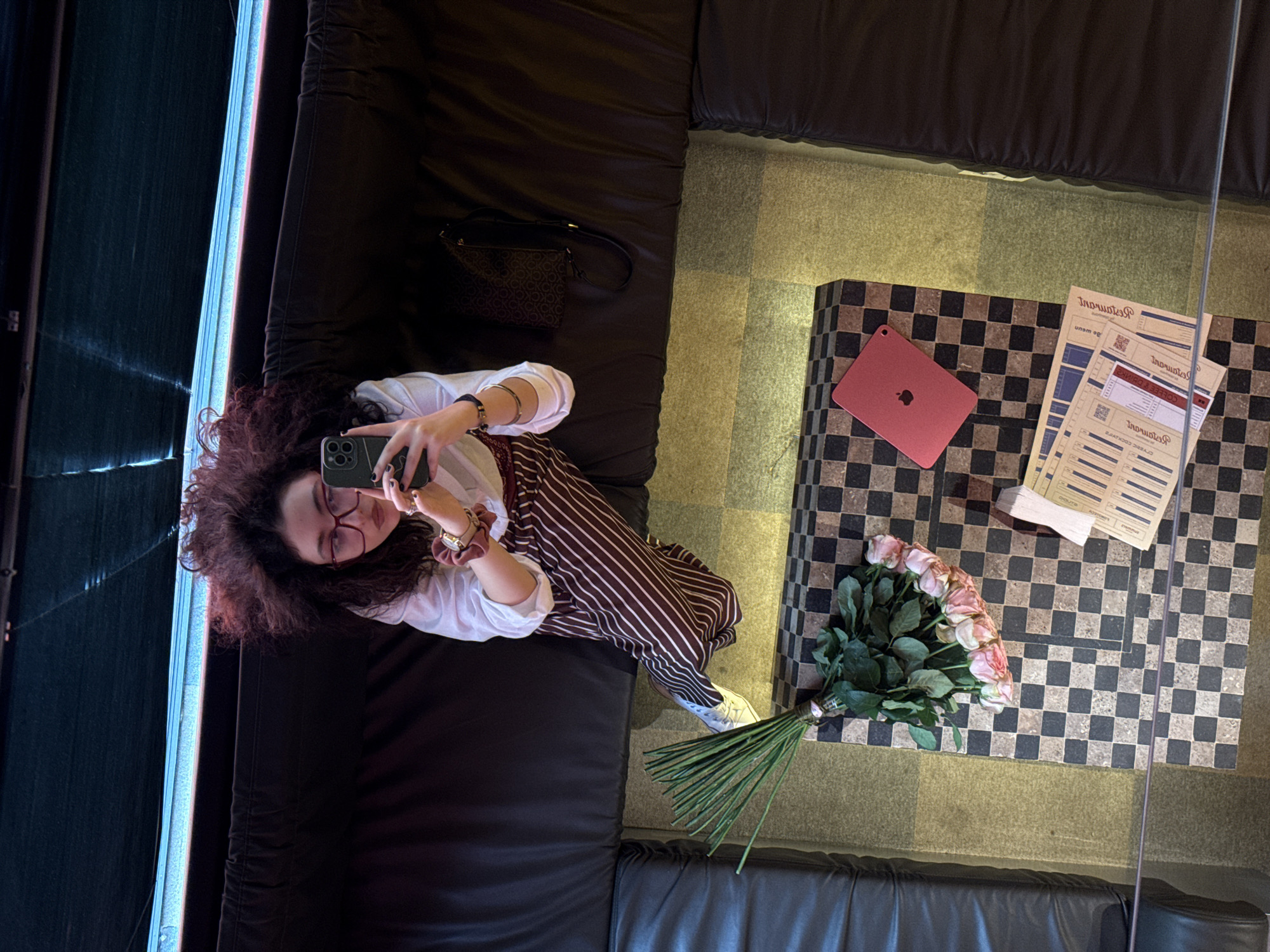 A woman with curly hair and glasses is taking a selfie with a smartphone, lying on a black leather couch. A small purse sits next to her. Next to the couch is a low table with a black and gold checkered pattern. On the table are a red tablet, a bouquet of pink roses, and several papers, presumably restaurant bills. The scene is in a restaurant in Yerevan, Armenia. The lighting suggests it's daytime, with light coming in through a window. The woman appears to be capturing the moment in the restaurant's lounge area.