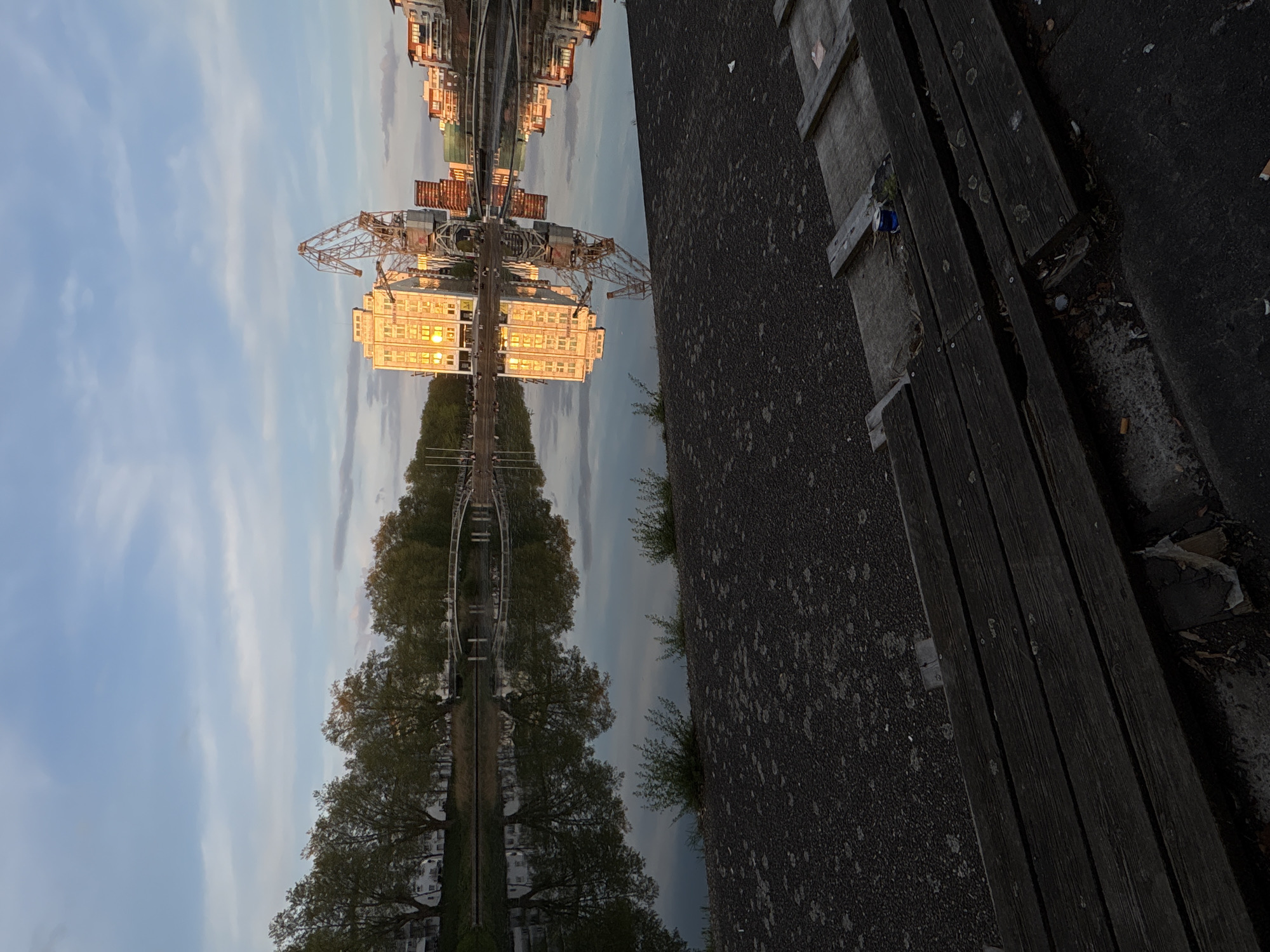 The image captures a reflection in the water in Strasbourg, France. The reflection shows a building with many windows catching the light, a tall crane structure, and trees lining a path. A stone or asphalt path runs along the right side of the image, with patches of vegetation growing along the edge. Adjacent to the path is a wooden structure, possibly a bench or part of a dock, with gaps between the planks. The reflection creates a symmetrical composition with the sky and clouds mirrored below. The image appears to be taken at either dawn or dusk, given the angle and color of the light. There are no people visible in the image.