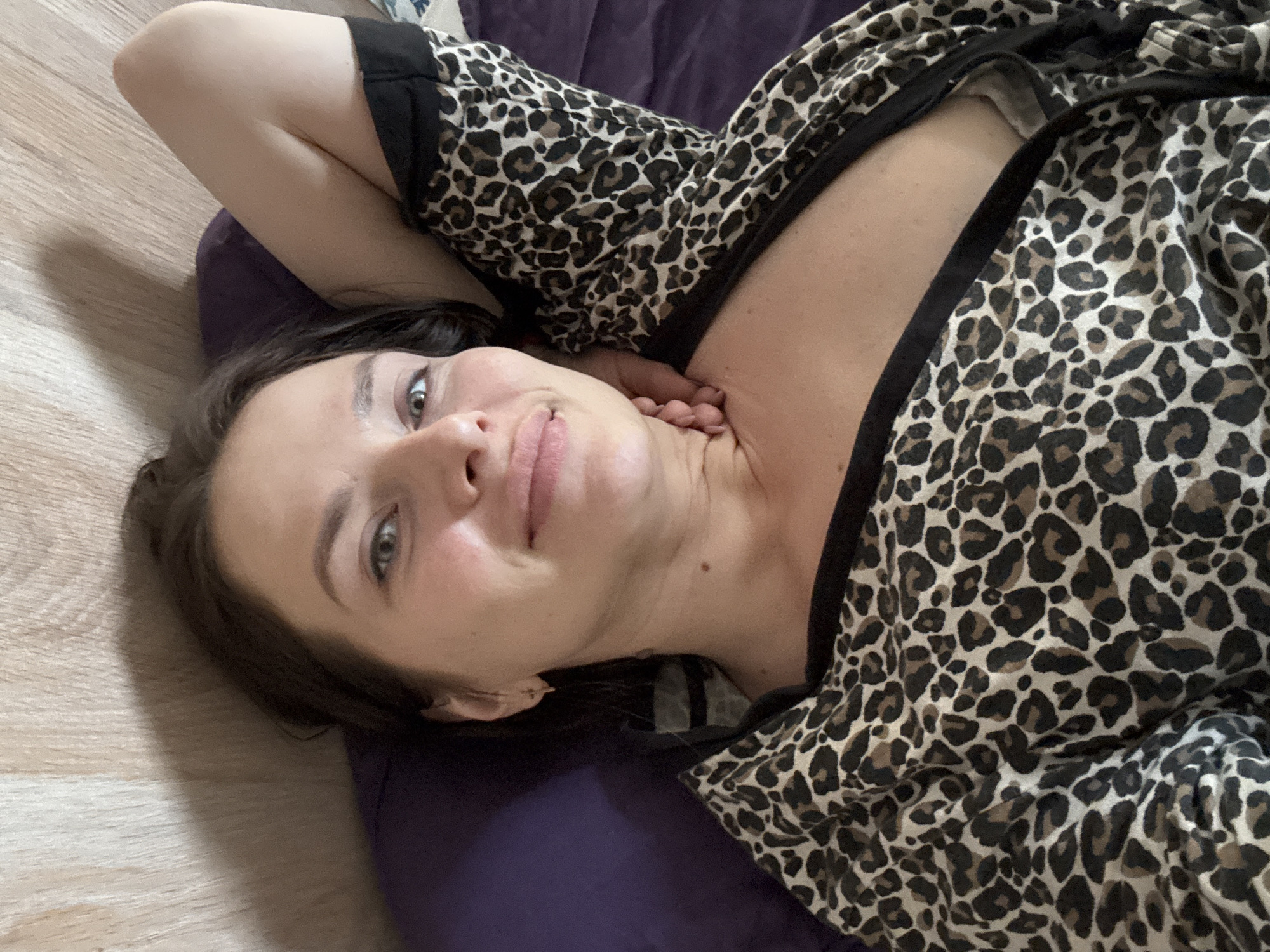 The image shows a woman lying on a purple cushion on a wooden floor. She is wearing a leopard-print top with black trim. Her right arm is raised above her head, and her left hand is near her neck. She is looking up at the camera with a slight smile. The location in the city of Rusalevka, Russia is not discernible from the image.