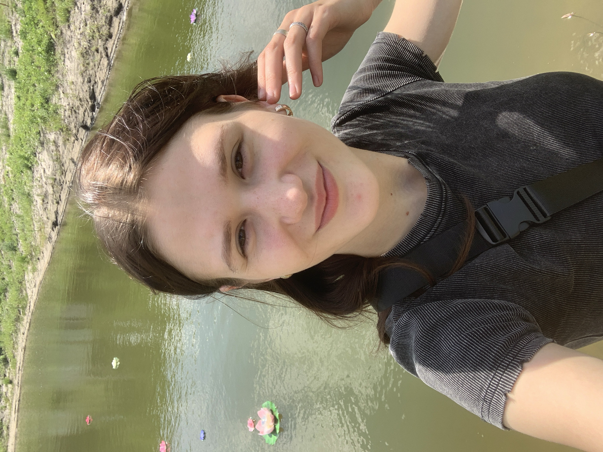 The media file is a selfie of a young woman taken outdoors during the day, likely in the late morning or early afternoon given the bright lighting and shadows. She is smiling and has her hand resting on her head, adorned with two rings. She is wearing a dark, ribbed t-shirt and a black fanny pack across her chest. The background shows a body of calm, green water, possibly a lake or pond, with several decorative objects resembling water lilies floating on the surface. The water is bordered by a muddy bank covered in green grass. The overall mood is cheerful and relaxed, suggesting a leisurely outing. The location is identified as Vasilyevka, Russia. There is no visible text in the image.