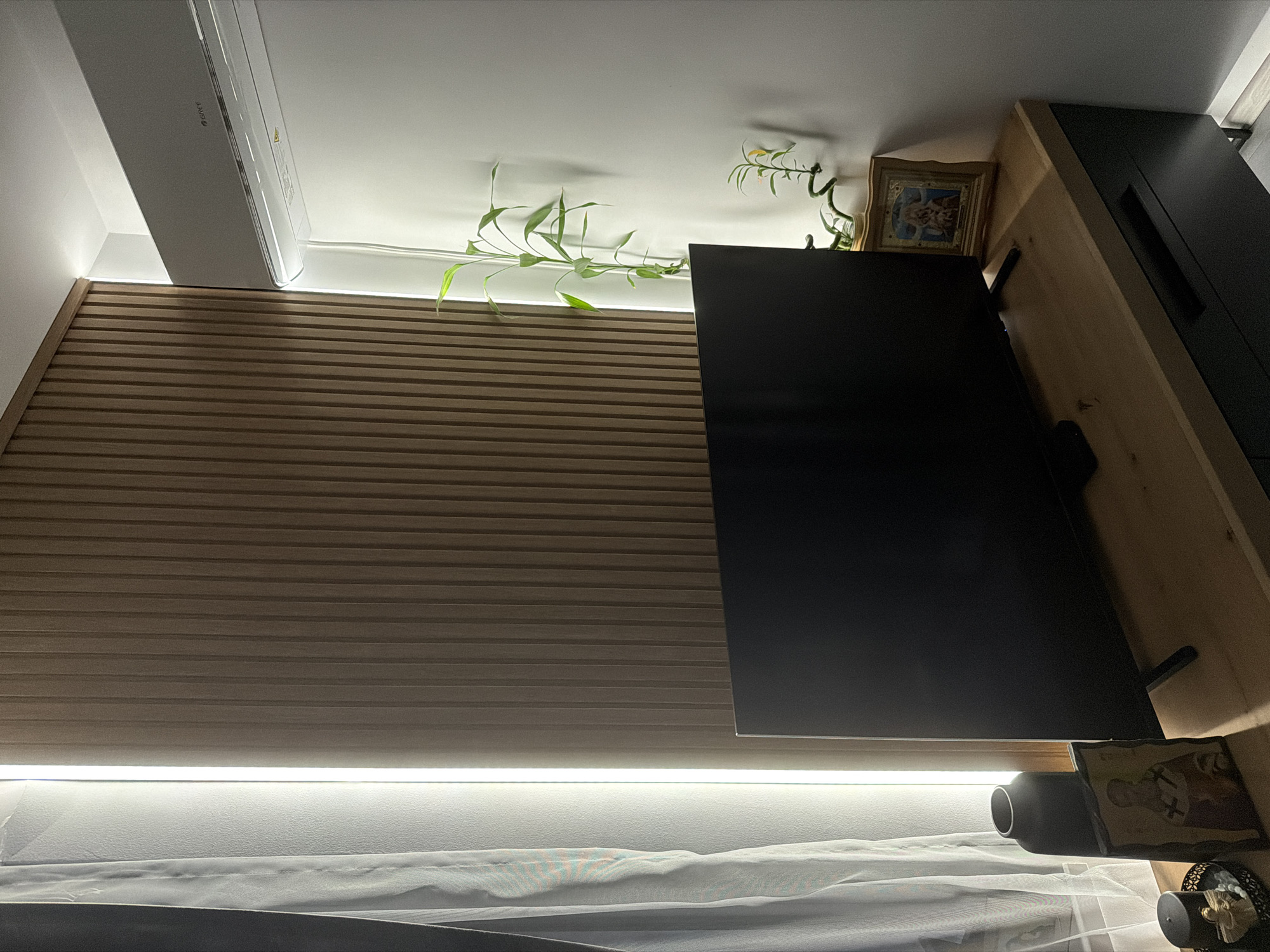 This media file captures an indoor scene, likely a living room or bedroom, in Burgas, Bulgaria, characterized by modern design elements and accent lighting, presumably at night or in the evening.
The left side of the room features a prominent wall covering made of light brown, horizontal wooden slats. Above this slatted wall, near the ceiling, a white air conditioning unit is installed, bearing the brand name "GREE". A bright LED strip light runs horizontally along the junction of the ceiling and the wall, illuminating the top part of the room and highlighting some green, leafy plants that appear to be trailing from the top edge of the slatted panel.
On the right side of the image, a black flat-screen television is positioned on a light-colored wooden shelf or media console. Above and slightly to the right of the television, a dark-colored cabinet with a vertical handle is visible. On the wooden shelf, next to and behind the television, decorative items include a small, framed religious icon/picture with a golden frame and a darker, perhaps older, icon or picture accompanied by a dark vase-like object.
Below the slatted wall and the television, another bright LED strip light runs horizontally, casting light downwards onto a white, sheer curtain, suggesting the presence of a window or balcony door. No specific activity or event is taking place; the image presents a static view of a well-lit and decorated interior space.