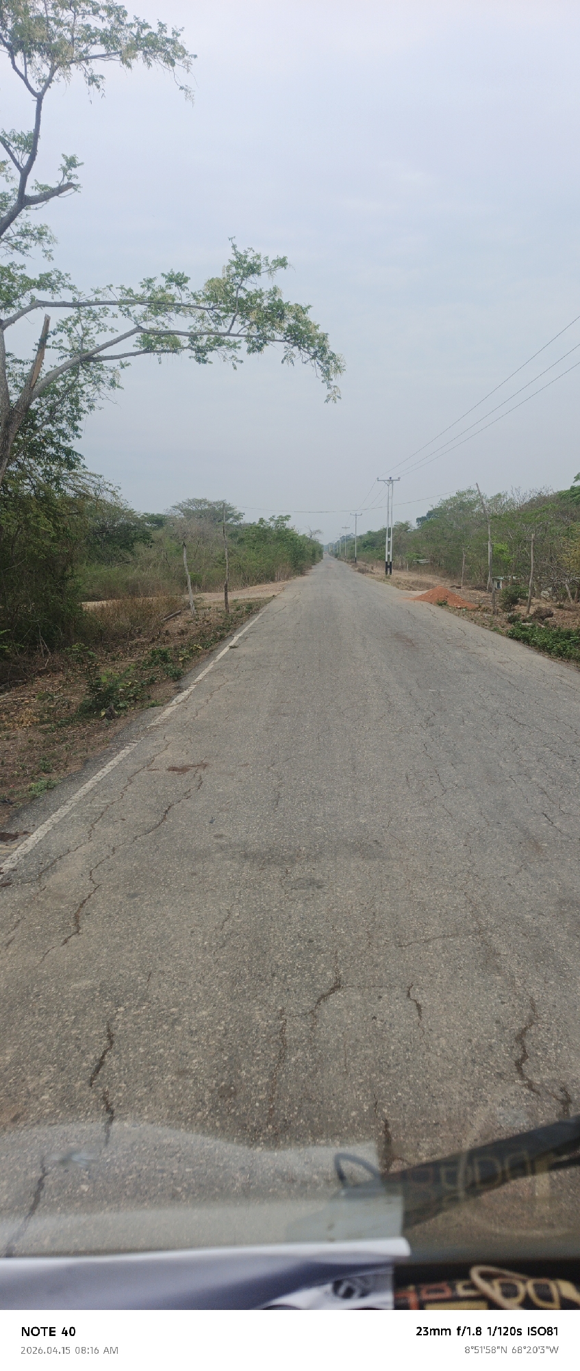 Here is a detailed description of the image:
**Setting:**
The image captures a long, straight paved road cutting through a rural landscape. The scene is set in El Baúl, Venezuela, as indicated by the GPS coordinates 8°51'58"N 68°20'3"W embedded in the photo data.
**Road and Surroundings:**
The road is gray, cracked, and shows signs of wear and tear. Along both sides of the road, there is dense vegetation consisting of trees and shrubbery. Utility poles line the road at intervals, with power lines visible above. A small pile of red dirt or rubble is present on the right side of the road.
**Sky and Light:**
The sky is overcast, providing soft, diffused light across the scene.
**Foreground Details:**
The image is taken from what appears to be the inside of a vehicle. The bottom portion of the image shows a blurry view of the dashboard or other interior elements, suggesting the photo was taken from a moving vehicle.
**Photo Data:**
The photo data at the bottom of the image indicates it was taken on April 15, 2026, at 08:16 AM, with a 23mm lens, f/1.8 aperture, 1/120 second shutter speed, and ISO 81.