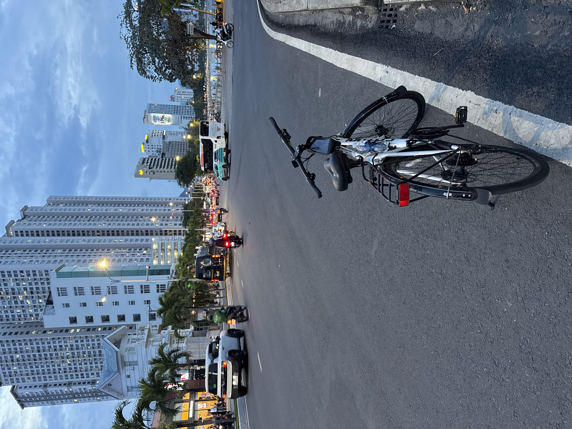The image depicts a street scene in Bac Nha Trang Ward, Vietnam, during what appears to be dusk. In the foreground, a white bicycle with black accents is parked on the side of the road, leaning against the curb. The bicycle has a water bottle attached to the frame and a red reflector on the rear fender. The road itself is asphalt with a white line marking the lane.
In the background, there is moderate traffic on the multi-lane road. Several vehicles are visible, including cars, motorcycles, and a bus. A white SUV is in the left lane, and a green car is in the right lane. A black van is also present in the traffic. Motorcycles are moving in both directions.
Towering buildings line the left side of the street, displaying numerous lit windows, indicating the city is urban and populated. Palm trees are interspersed along the sidewalk. On the right side of the street, beyond the road and a pedestrian walkway, a body of water can be seen, with a tree and more distant buildings along its shore. People are visible walking along the waterfront area. The sky is overcast with a twilight hue.
