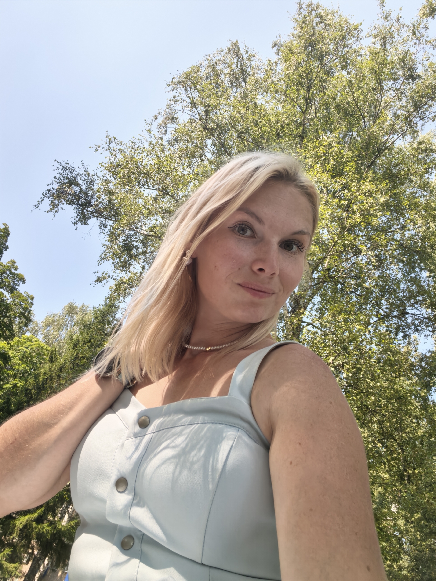 A blonde woman is taking a selfie outdoors. She is wearing a light blue dress with buttons down the front and a pearl necklace. Her left arm is raised, and her hand is near her hair. The background is a tree with green leaves and a clear blue sky. The image is an outdoor shot taken during the day.