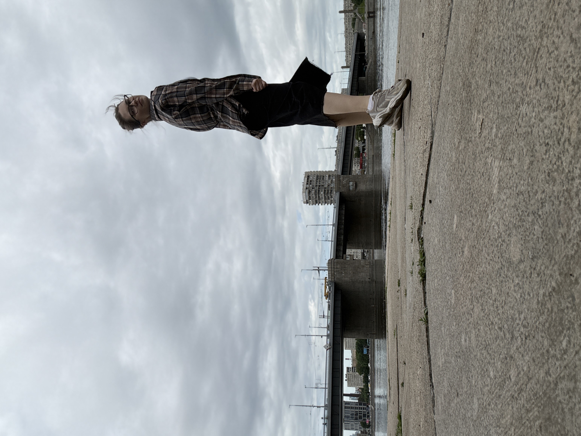 A young woman stands on a concrete embankment in an urban environment. She is viewed from a low angle, with the camera positioned on the ground in front of her. The woman is wearing a plaid shirt, black skirt, and athletic shoes. Her hair is slightly windswept, and she is looking over her shoulder at the camera.
In the background, a large bridge spans a body of water, with city buildings visible beyond it. The sky is overcast with clouds, suggesting a cool or temperate climate. The concrete embankment is cracked and weathered, with patches of dry grass growing through. The scene appears to be part of a city park or waterfront area, common in many urban landscapes. The overall impression is one of a casual moment captured outdoors.