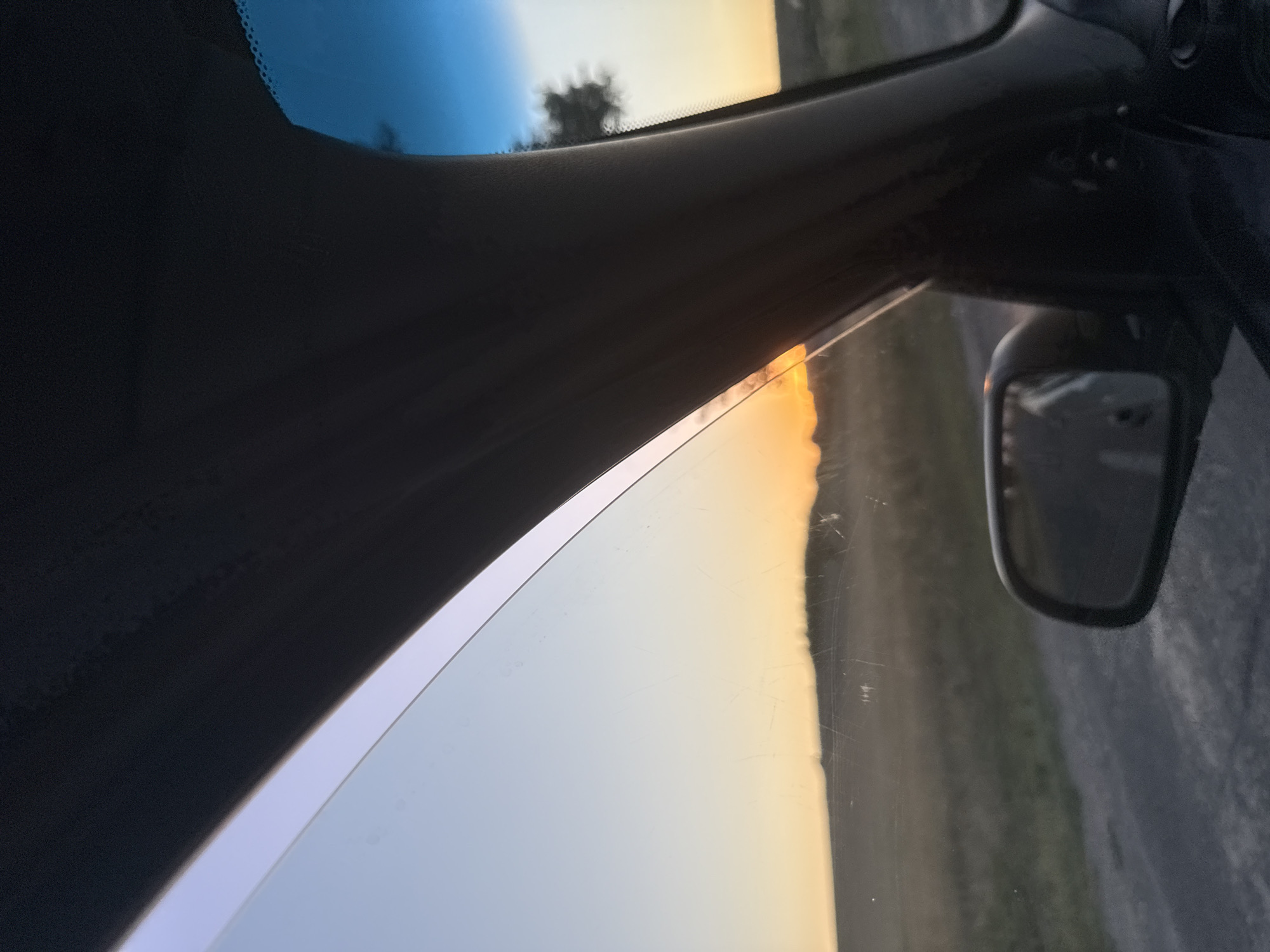 The image captures a view from inside a vehicle, looking outwards towards a sunset. A portion of the car's interior, specifically the A-pillar and the top of the windshield, frames the scene. The sky transitions from a pale blue at the top to a warm orange and yellow hue near the horizon, indicating it is either sunrise or sunset. A landscape of rolling hills or fields is visible beneath the sky, with a road running in the foreground. A side-view mirror is present on the right side of the frame, reflecting a blurred image. A solitary tree is silhouetted against the sky in the distance. The overall impression is one of a rural or semi-rural setting in Cioc-Maidan, Moldova, during twilight.